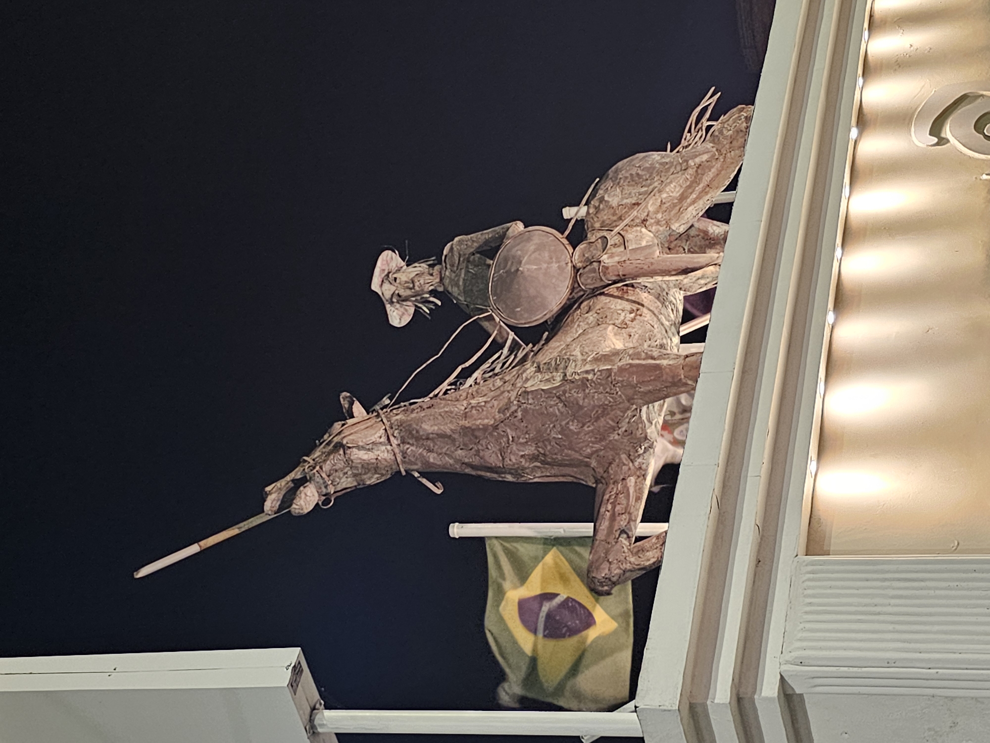 The image is a low-angle shot of a weathered, metallic sculpture of a man on horseback, wielding a lance, positioned on a building facade in Armação Dos Búzios, Brazil. The Brazilian flag is visible beneath the sculpture, attached to the building. The dark night sky forms the backdrop. The building's architectural details, including lit architectural molding, are on the right of the image. The horse and rider are the primary focus, with the flag and building details providing context.