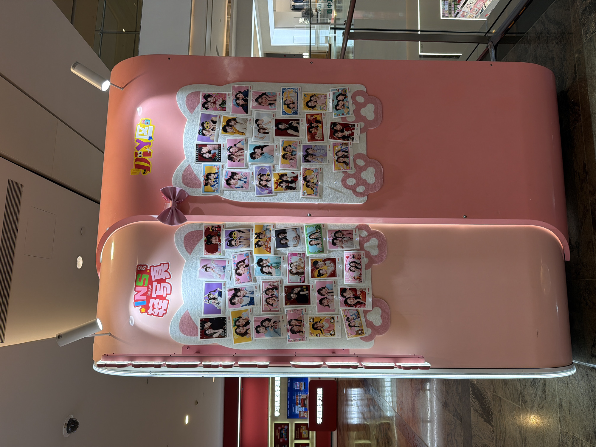 This media file captures two stacked, bubblegum-pink photo booths or kiosks situated in an indoor commercial setting, likely a shopping mall in Yuzhong District, China. The booths have rounded edges and feature decorative white felt shapes resembling cat ears and paw prints, along with a pink bow on the upper unit.
Both booths display numerous small, printed photographs, reminiscent of instant prints or photo strips, arranged on their felt surfaces. These photos predominantly show young people, primarily females, posing in pairs or small groups. Their cheerful expressions and playful interactions suggest a fun and engaging experience. Spotlights extend from the top of each booth, illuminating the displays.
Visible text on the upper booth includes "DIY" and "COLOR ROBO". On the lower booth, Chinese characters read "学而思学习机" (Xueersi Learning Machine) and "轻写真" (Light Photo/Portrait), accompanied by "INS" and "COLOR ROBO," with "SHAILE" also partially visible. In the background, reflections on the polished floor and blurry signage with Chinese characters like "学习机就选" and "中工" are discernible.
The bright, even lighting indicates an indoor setting, and the modern design of the booths targets a youthful demographic. The overall scene implies a vibrant atmosphere where people can create and share personalized photo memories.