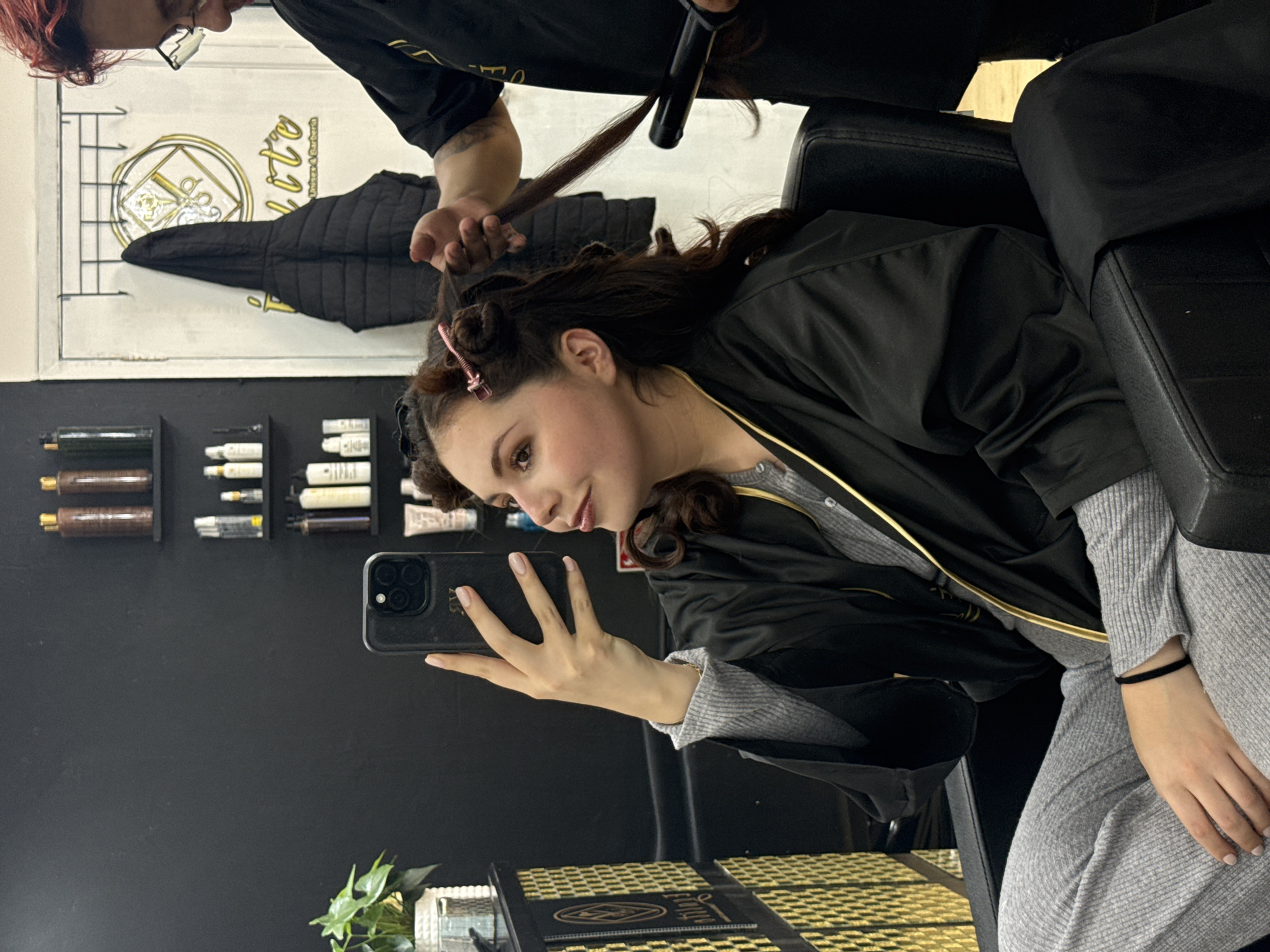 A woman is seated in a black salon chair, taking a selfie with her smartphone. Her hair is being styled with a curling iron by another individual. The stylist is wearing a black shirt. The woman has clips in her hair, and is wearing a grey outfit with a black jacket draped over her shoulders. The salon has a black wall with floating shelves holding various hair care products. A logo is visible in the background, along with a black jacket hanging on a coat rack. A plant sits on the floor. The image appears to have been taken inside a salon in Mexico City, Mexico.