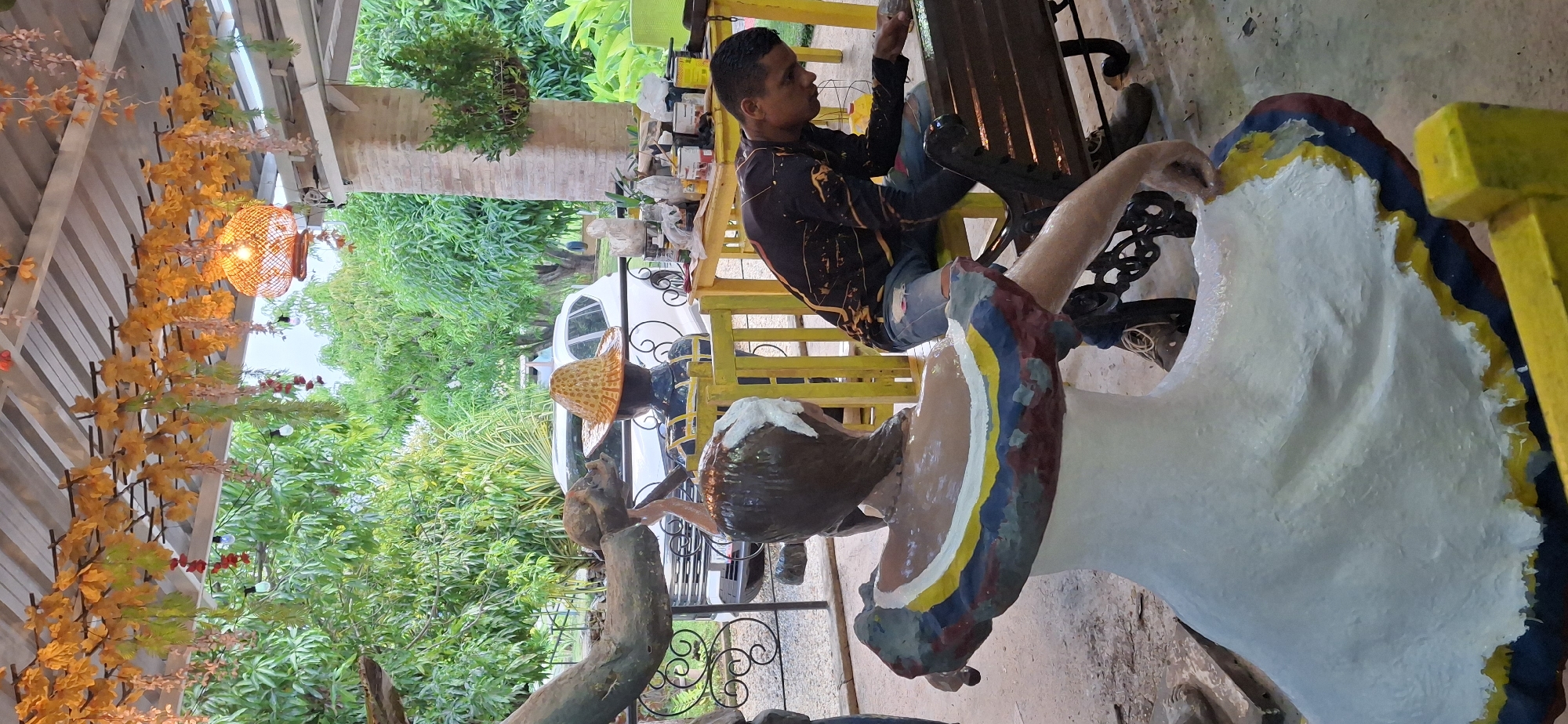 In Vallecito, Venezuela, a man is meticulously painting a sculpture of a woman. The sculpture, with its unfinished white dress adorned with stripes of blue, yellow, and red, stands in the foreground. The woman's back is turned to the viewer, and her hair is pulled back. The man, wearing a black long-sleeved shirt with a paint-splattered design and ripped blue jeans, is seated on a dark metal bench, focused on his work. He holds a brush in his right hand and appears to be applying paint to the bench itself, or possibly touching up the sculpture from a closer angle. The scene is outdoors, beneath a covered structure decorated with artificial fall foliage and hanging lights. In the background, lush green trees and foliage are visible, along with a white vehicle parked nearby. A yellow table, laden with various containers of paint and supplies, is situated behind the sculptures. The overall impression is one of creative activity taking place in a tropical, rustic setting.