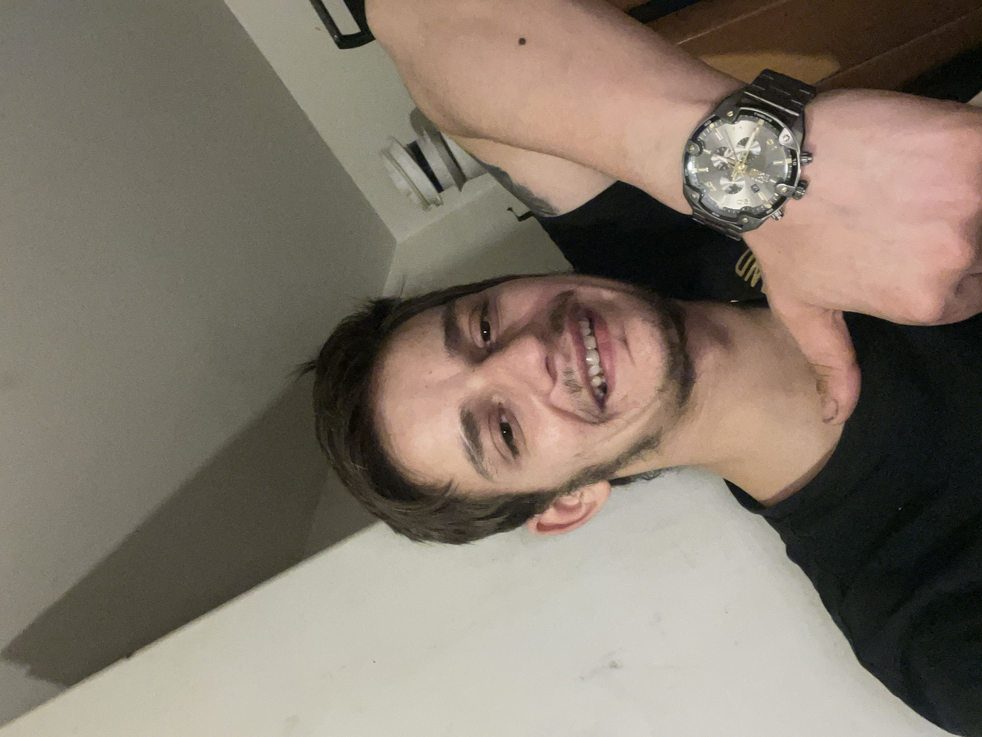 A man is lying on a white surface, possibly a bed or floor, with a white wall behind him. He is smiling and has short brown hair and a light beard. He is wearing a black T-shirt and a silver watch on his left wrist. There is a vent on the white wall behind him. There is no indication of the location in the city of Unknown, Unknown.