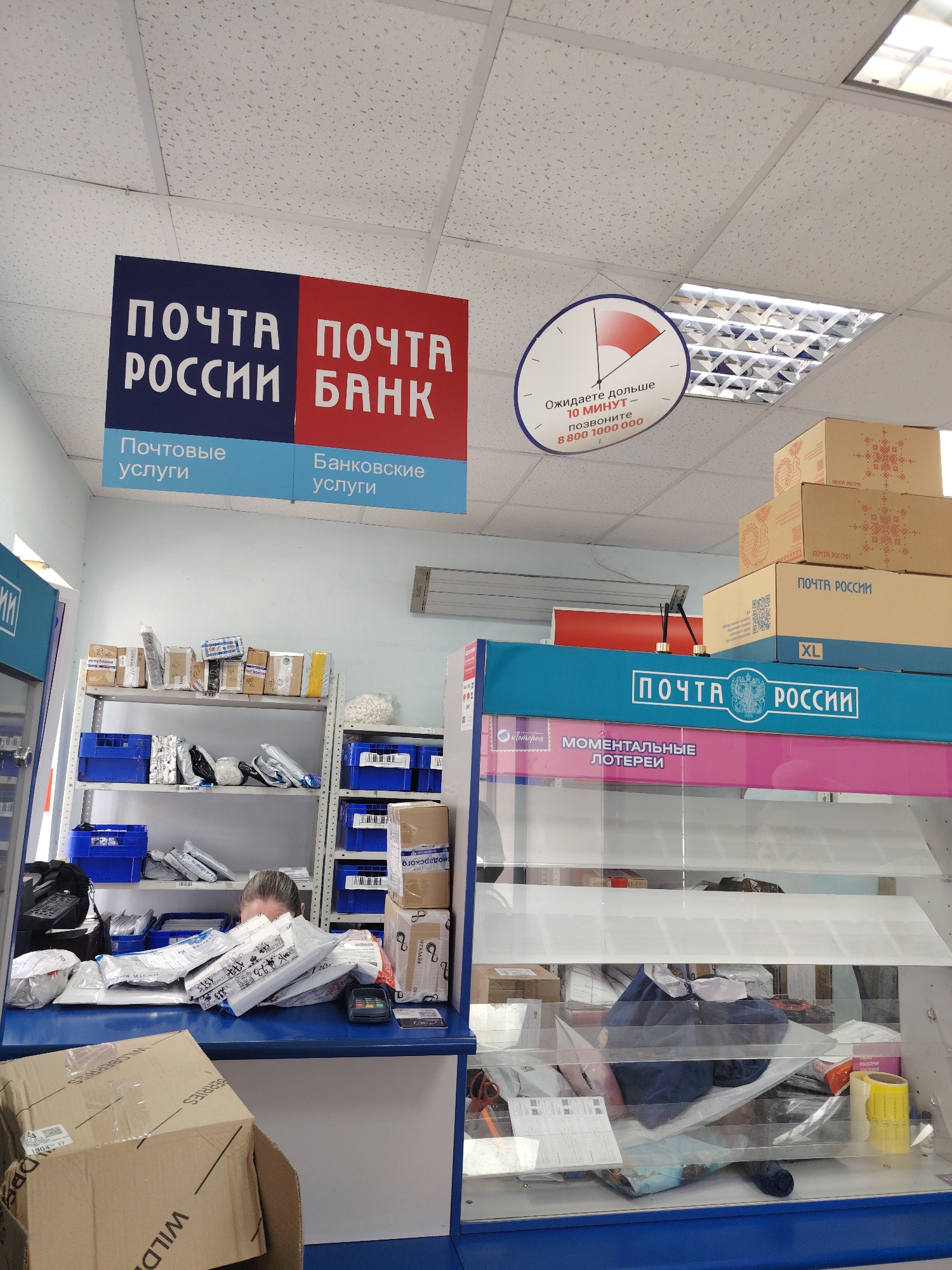 The image depicts the interior of a "Pochta Rossii" (Russian Post) and "Pochta Bank" branch. In the foreground, shelves are stacked with numerous packages and mail items. A person, partially visible behind a counter laden with envelopes and packages, appears to be an employee. The counter also has a payment terminal. Above, signs indicate "Pochta Rossii" and "Pochta Bank" along with services like "Post Services" and "Banking Services." A clock graphic on the ceiling suggests a waiting time of "over 10 minutes." Cardboard boxes, some labeled "Pochta Rossii," are stacked to the right. The environment appears to be a typical postal service and banking facility. While the image is taken in a Russian Post and Bank, there is no information within the image to confirm its location in Rostov-on-Don, Russia.