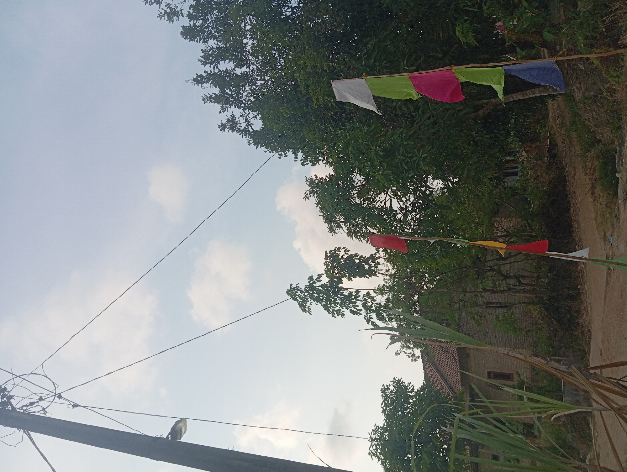 The image depicts an outdoor scene with a prominent utility pole on the left, adorned with electrical wires and a street lamp. In the foreground, there are several colorful flags suspended from poles, including white, green, pink, red, and blue. Behind the flags, lush green trees dominate the midground, interspersed with a house with a tiled roof and brick walls. The sky above is partly cloudy. The overall impression is of a rural or suburban setting. The presence of flags suggests a festive occasion or cultural event. The location is identified as Dente Teladas, Indonesia, which is consistent with the tropical foliage and architectural style of the house.