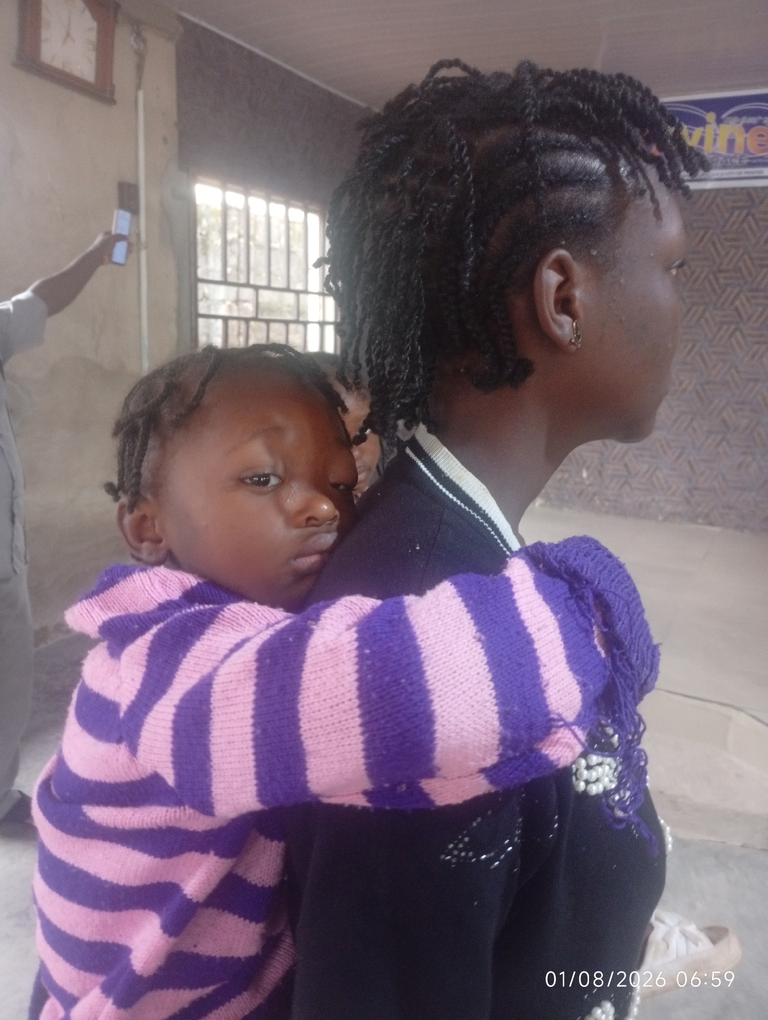 A young Black child, with braids and wearing a pink and purple striped sweater, is carried on the back of a Black woman, who is also wearing braids and a dark sweater. The woman is looking to her right, away from the camera. The child is looking at the camera with a serious expression. The background appears to be indoors, with a clock visible on a wall and a window with bars. A partial view of another person holding a phone is visible in the left side of the image. The overall lighting suggests an indoor setting. The date stamp at the bottom right indicates August 1, 2026, at 06:59.