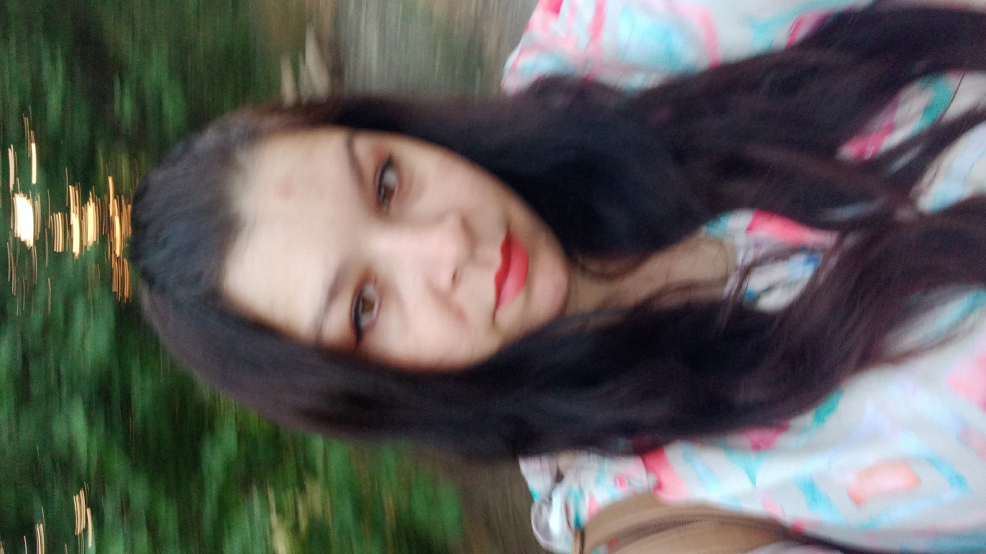 A woman is taking a selfie outdoors. She has long, dark hair and is wearing a colorful, patterned shirt. Her lips are painted red. In the background, there are blurred green trees and lights. The image is slightly out of focus due to motion blur. The location is not specified in the image or the provided context.