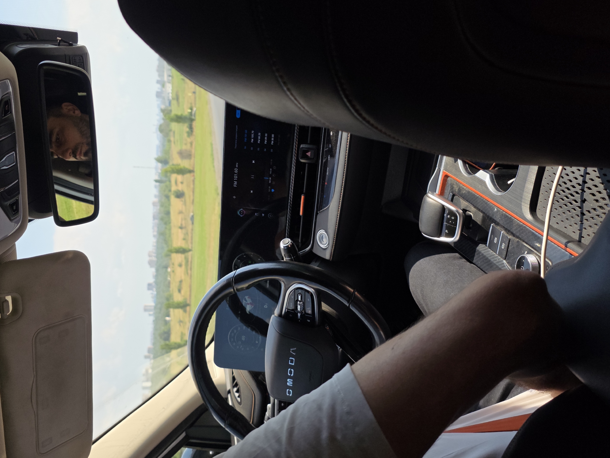The image shows the interior of a car with a view of the road ahead and a distant cityscape. A man is reflected in the rearview mirror, looking forward. The dashboard features a touchscreen display and a steering wheel with buttons. The driver's arm is visible on the steering wheel, and their hand rests near the gear shift. The car appears to be moving on a road surrounded by greenery, with trees and fields visible in the background. The cityscape in the distance suggests an urban environment. Based on the visual cues, the location is identified as Rostov-on-Don, Russia, possibly on a road leading into the city with a view of the Don River and its embankment.