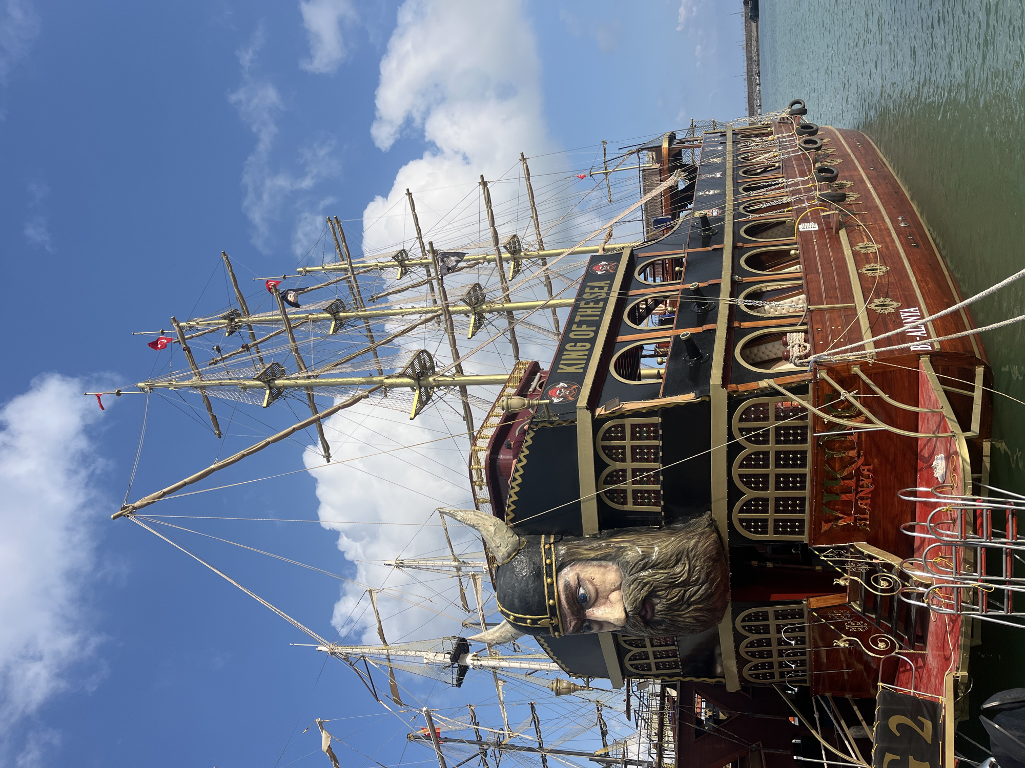 The image shows a large, ornate ship docked in what appears to be the city of Alanya, Turkey, based on the writing "B-Alanya" on the ship's stern. The ship is elaborately decorated, with a figurehead resembling a Viking with horned helmet at the bow. The hull is painted in brown and black, with gold trim. The name "KING OF THE SEA" is written in large letters on the side of the ship. It has multiple masts and rigging typical of a sailing vessel. The sky is blue with scattered white clouds. The water next to the ship appears green. There are no people visible in the image. The ship is docked at a pier.