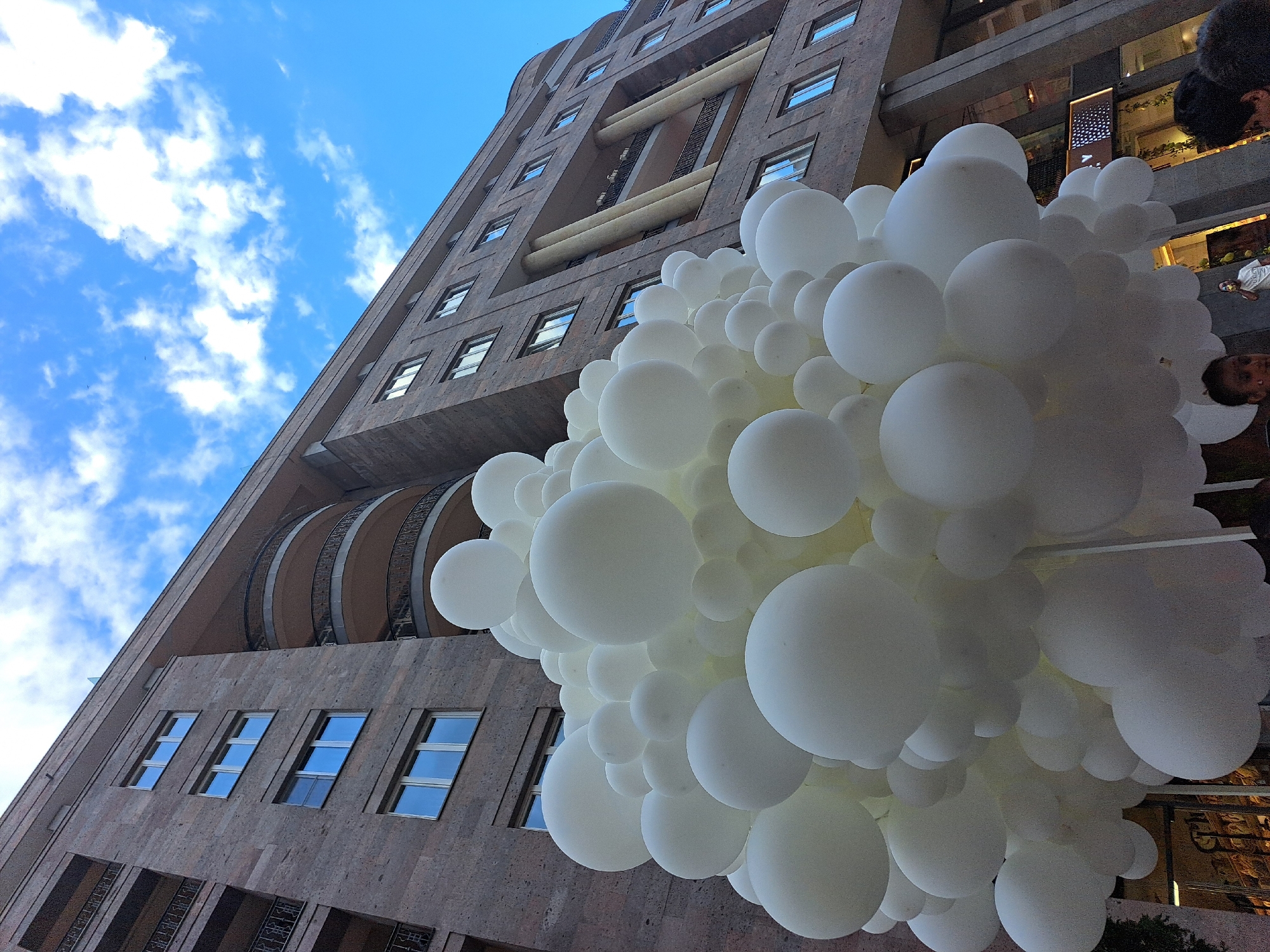 The image shows a tall, multi-story building in Yerevan, Armenia, partially obscured by a large cluster of white balloons. The building has a textured, brown exterior with multiple windows and distinctive architectural details, including recessed sections and curved balconies. The sky above is blue with scattered white clouds. In the foreground, the white balloons dominate the view, creating a cloud-like effect. Parts of people can be seen behind the balloons on the right side. The scene suggests a festive or celebratory event in an urban setting.