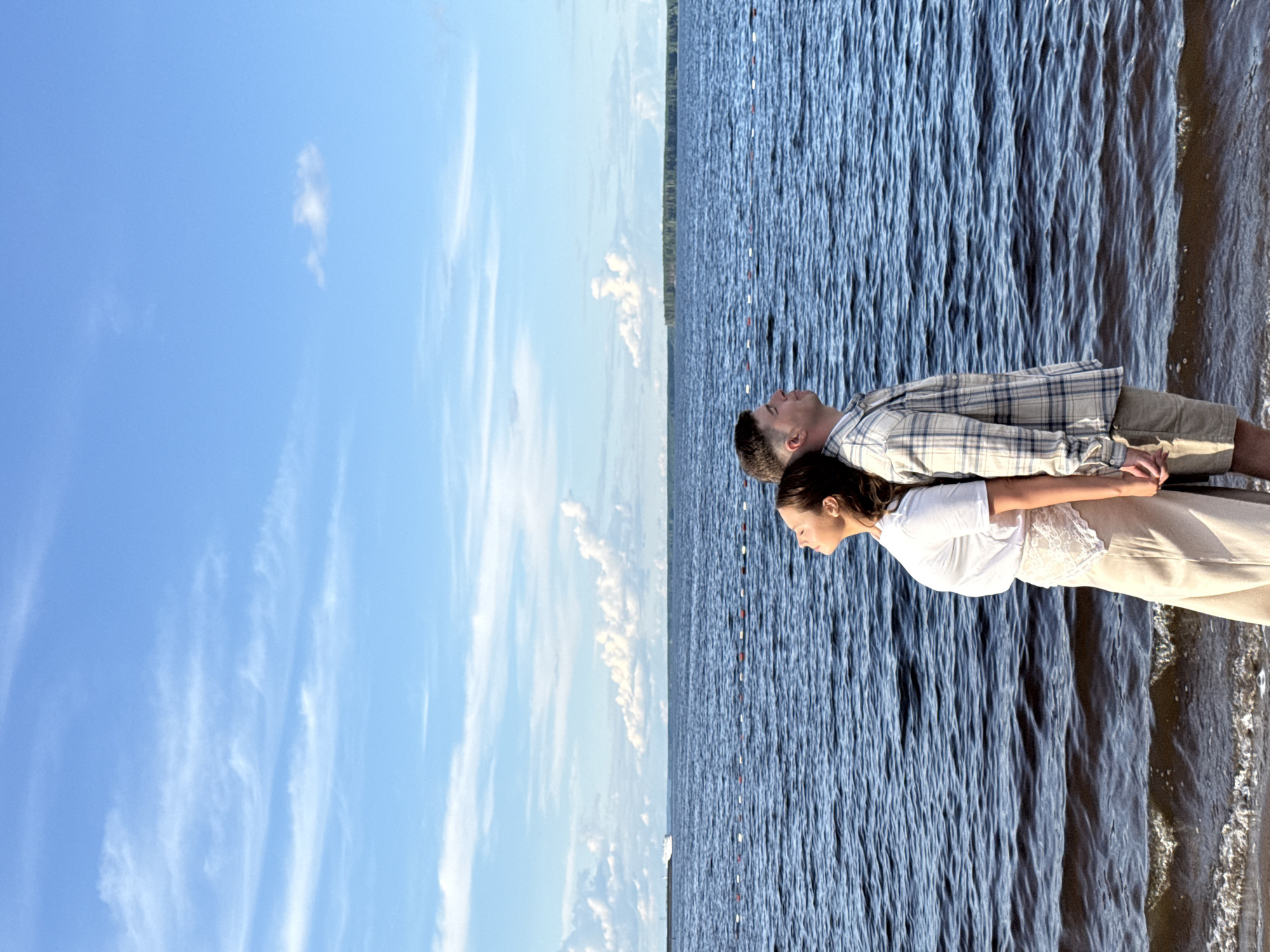 A couple is lying side-by-side on what appears to be a dock or edge of a body of water, with a backdrop of sky and water. The image is rotated 90 degrees counter-clockwise. The man is wearing a plaid shirt and shorts, and the woman is wearing a white top and light-colored pants. They are positioned close together, their heads near the center of the frame, and appear to be relaxing. To the left is a bright blue sky with wispy clouds. To the right is a dark blue body of water with small waves and the shoreline visible. The location is likely the city of Chaikovsky, Russia, as specified in the prompt.