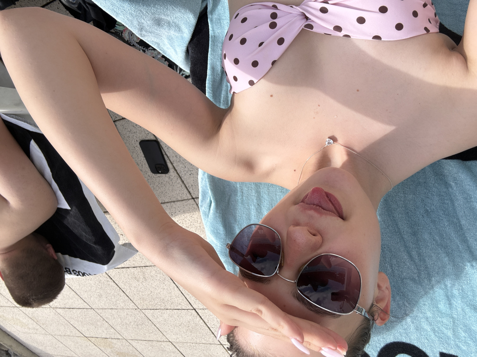 The media file shows a young woman lying on her back on a light blue towel, outdoors in what appears to be a sunny environment. She is wearing a pink and brown polka dot bikini top and sunglasses. Her left hand is raised, partially covering her face. A thin silver necklace with a small pendant is visible around her neck. Her tongue is sticking out slightly. To her left, a black smartphone is lying on a tiled surface. Further to her left, partially visible, is the head and upper torso of another person lying down. The background consists of a tiled patio and what looks like the edge of a swimming pool. The lighting suggests it is daytime, likely sunny, as indicated by the shadows and highlights. The overall scene conveys a relaxed, summery atmosphere, possibly at a swimming pool or by a body of water. No visible text is present in the image.