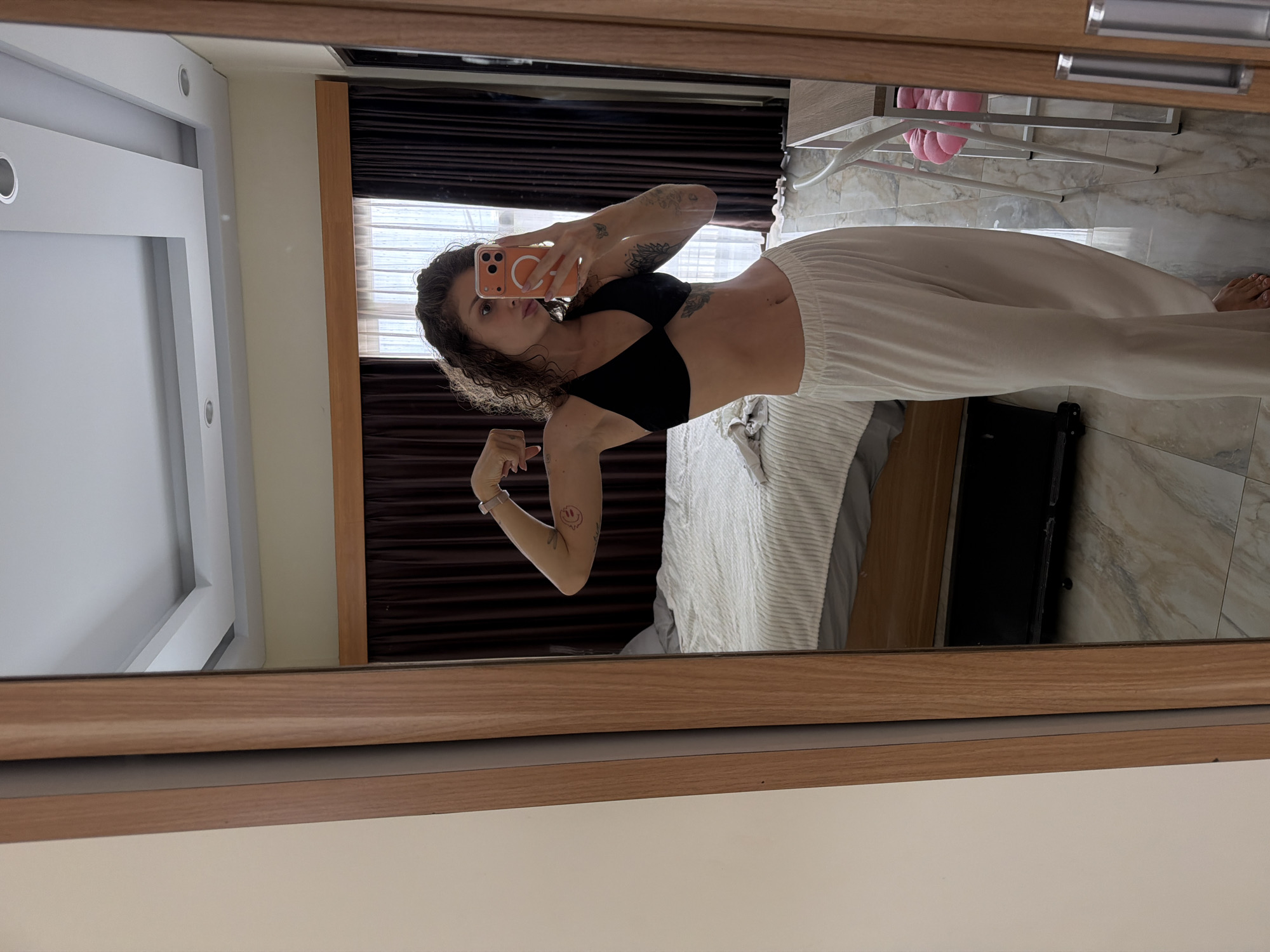 An individual with curly brown hair, wearing a black sports bra and white pajama pants, is taking a mirror selfie in a bedroom. The person is flexing their bicep and holding an orange phone. Tattoos are visible on the arms and torso. The room features a bed with white bedding, dark brown curtains, and a tiled floor. A wooden-framed wardrobe or door is to the left, and part of a desk and chair are visible in the reflection. The environment suggests an indoor residential setting in North Kuta, Indonesia.