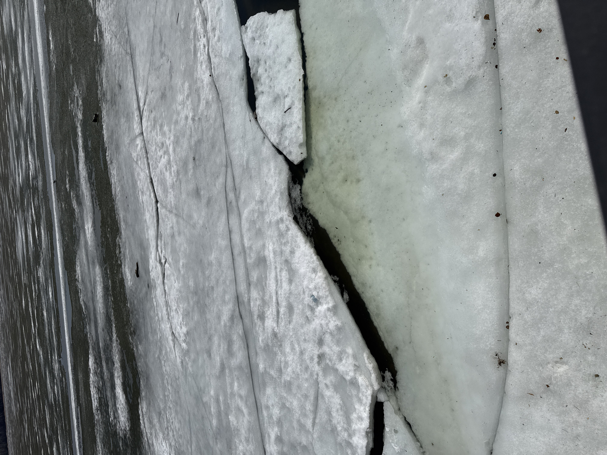 The image shows a close-up view of broken ice, likely on a body of water. There is a dark, narrow gap between the ice fragments, potentially revealing water underneath. The ice itself appears textured, with variations in color and small debris scattered on the surface. The left side of the image shows a darker, possibly melted section, indicating a variation in thickness or melting patterns. The scene is presumably located in or near Chaikovsky, Russia, but there are no identifiable landmarks or objects to confirm this directly.