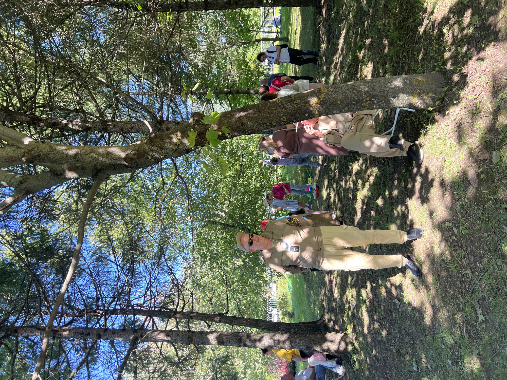 A group of people are gathered in a park on a sunny day. The foreground is dominated by a large tree and the lower body of a man standing in a khaki uniform and cap. He is wearing sunglasses and has a badge clipped to his shirt. Behind him, other people are visible, including a woman in a yellow shirt, children in casual clothing, and adults in various attire. The background shows more trees, foliage, and some structures that appear to be part of a park or recreational area. The ground is covered with grass and dappled sunlight. The scene suggests a casual outdoor gathering or event.