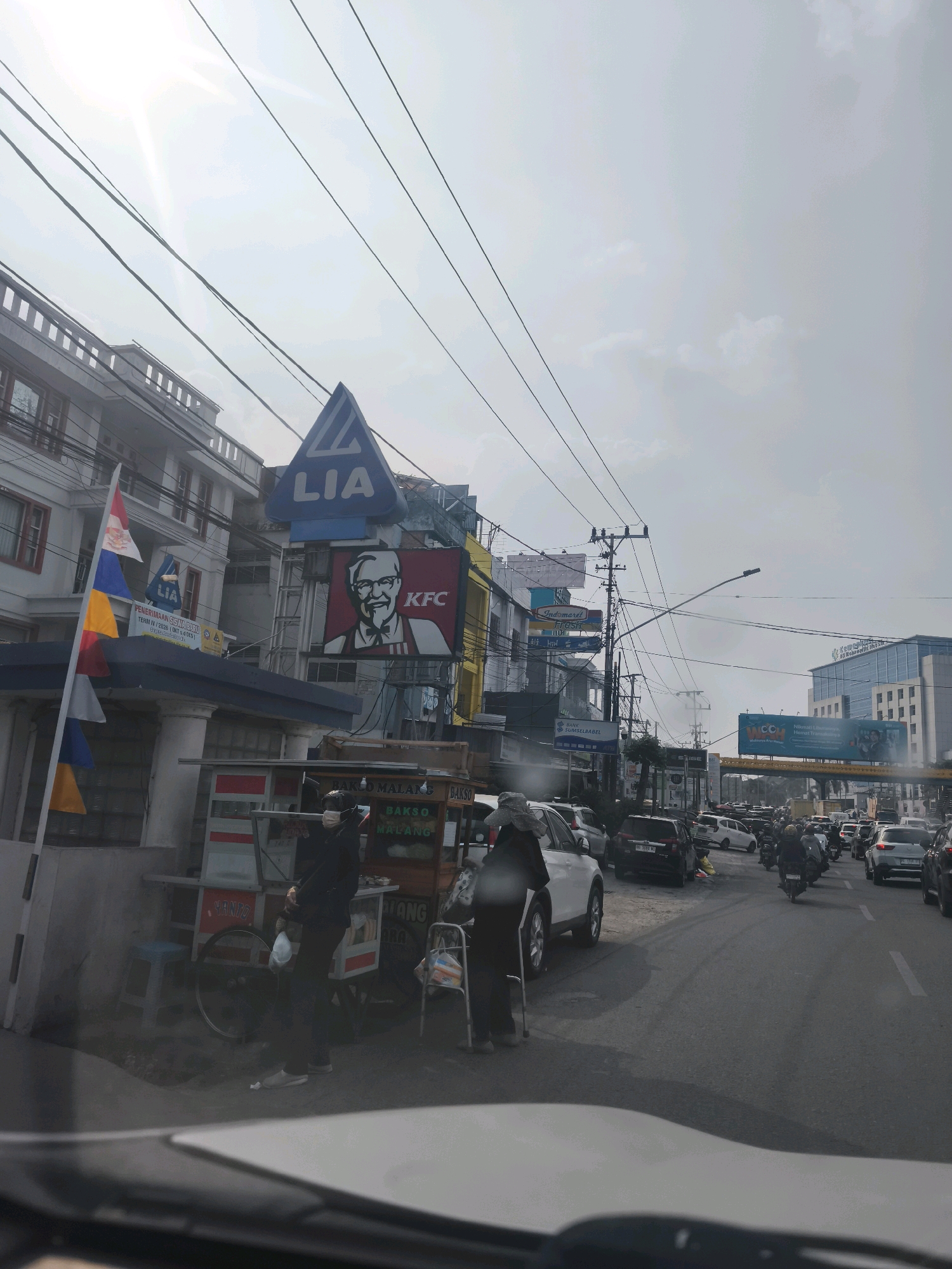 The image shows a street scene in Palembang, Indonesia, with a clear sky and visible power lines overhead. On the left side of the street, there is a multi-story building with shops on the ground floor. A prominent blue triangular sign with "LIA" in white letters is mounted above a KFC restaurant with the iconic Colonel Sanders logo. In front of the shops, a food cart selling "Bakso Malang" is visible, with two people standing near it, one of whom appears to be a vendor wearing a mask. Colorful flags are also displayed on a pole.
The street is busy with traffic, including cars, motorcycles, and a pedestrian on the left side of the road using a walking aid. Several vehicles are parked along the roadside. In the background, taller modern buildings are visible, including one with a blue glass facade that appears to be a commercial or office building. A large billboard with text and images is also present in the distance. The overall impression is of a bustling urban environment on a bright day.