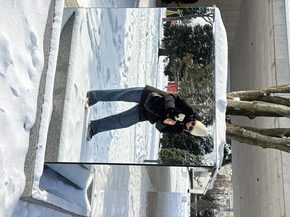 The scene depicts a snow-covered urban park in Plodorodny, Russia, on a bright winter day. A rectangular art installation featuring mirrored sides rests on a grey granite base, both partially covered with snow. A female individual is captured in the mirror's reflection, positioned centrally. She is wearing a cream knit beanie, dark sunglasses, a black puffer jacket, black gloves, blue denim jeans, and athletic shoes (one light-colored, one dark). She holds a white smartphone horizontally with both hands, angled as if recording or photographing herself. A black crossbody bag is worn over her left shoulder. The background, visible in the mirror's reflection and directly beyond the structure, consists of snow-dusted bare deciduous trees, dark evergreen trees, and distant multi-story residential buildings. A yellow illuminated angelic figure and a red building are also visible in the reflected background to the left. The ground surrounding the installation is uniformly covered in snow, with footprints visible on the granite base.