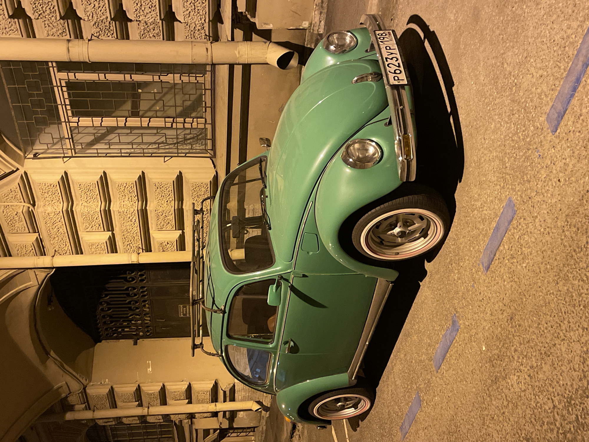 The image shows a vintage green Volkswagen Beetle parked on a street in Saint Petersburg, Russia. The car is parked on the right side of the frame and is oriented diagonally. The vehicle's license plate reads "P623YP 198 RUS". The street is marked with blue stripes, indicating parking lines. The background features an ornate building facade with barred windows, arched entrances, and decorative architectural details. The lighting suggests it is nighttime. There is no visible evidence of other people.