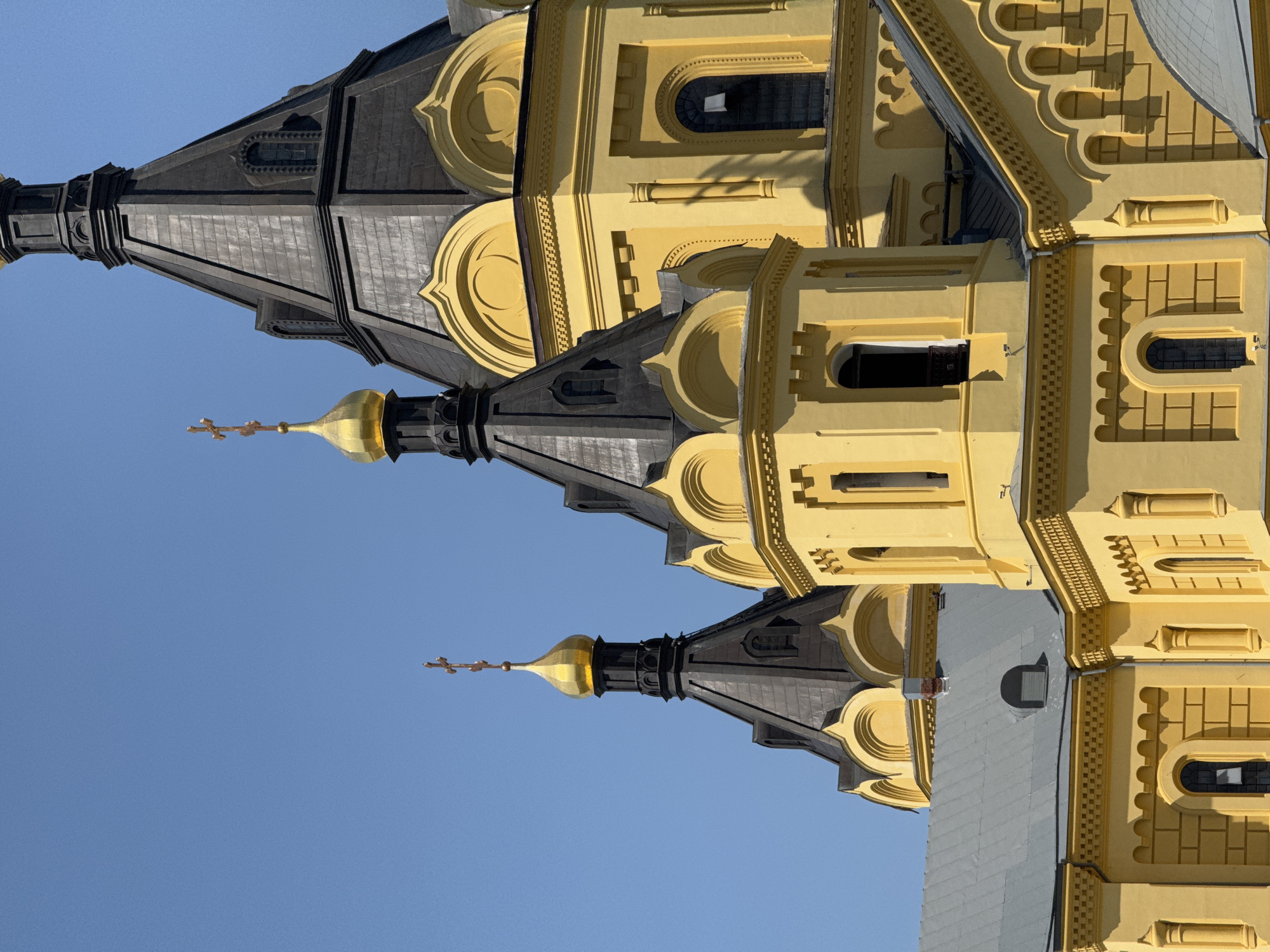 The image depicts the towers and domes of the Alexander Nevsky Cathedral in Nizhny Novgorod, Russia. The architectural elements are showcased against a clear blue sky. The towers exhibit a combination of yellow and dark gray coloring, with golden domes surmounted by crosses. The image is taken from a low angle, emphasizing the upward perspective of the cathedral's structure.