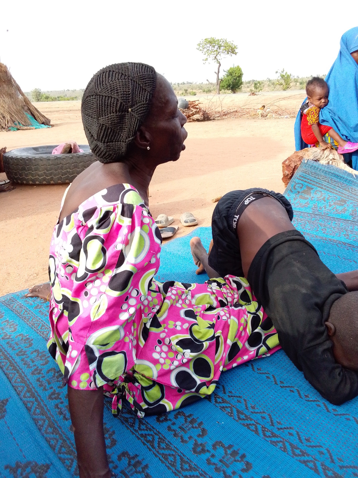 Location: Mulke Barikin Sayejo, Nigeria.
Scene: An outdoor setting. An elderly woman with dark skin sits on a blue patterned mat wearing a dress with pink, green, and black floral patterns and a black knit cap. A child wearing black shorts and a black shirt is lying face-down on the same mat. Another child is being held by a woman wearing a blue head covering. A stack of firewood is present in the background. A large tire rests near a straw hut.