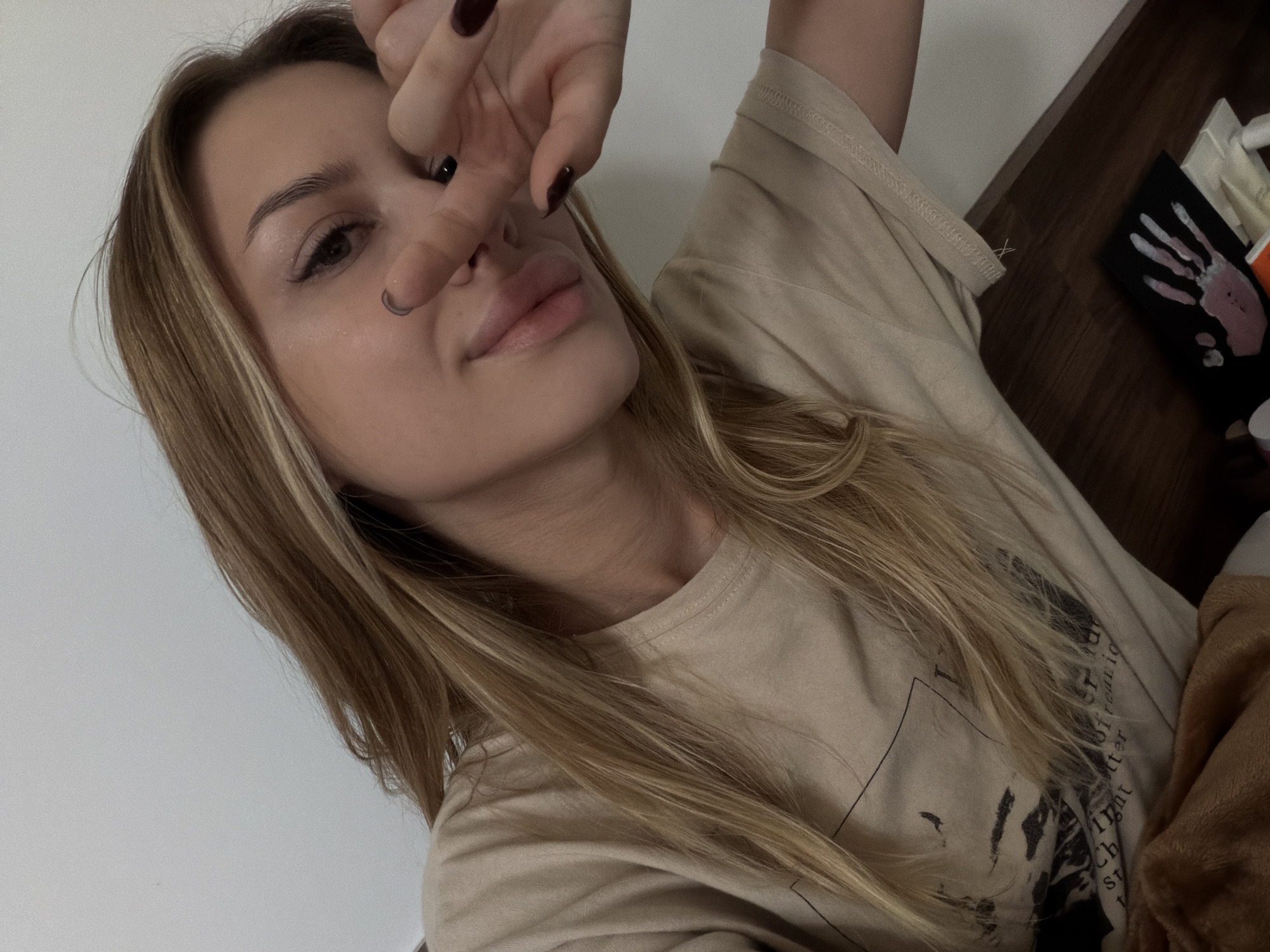 The image shows a young woman with blonde hair and light skin, wearing a tan t-shirt. She is looking at the camera with her hand covering part of her face, making a peace sign. Her lips are slightly parted, and she has on dark nail polish. The background is a plain, light-colored wall, and a wooden surface is visible at the bottom. There is a blanket or soft fabric in the lower left corner, and a black canvas with a pink and white handprint on it is on the wooden surface. The scene appears to be indoors, in a casual setting. There is no indication of the city of Đà Nẵng, Vietnam.