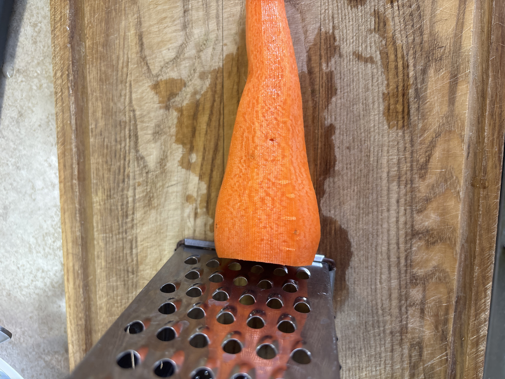A close-up shot captures a portion of a peeled carrot positioned on top of a metal grater. The carrot, a vibrant orange, tapers to a point and sits vertically, its textured surface visible. Below it, the grater's flat surface is dotted with numerous circular holes, some of which reveal the carrot's orange hue through them. The grater itself appears to be made of stainless steel and has a slightly worn appearance.
The backdrop is a wooden cutting board with a prominent grain pattern. The wood is a warm, light brown, marked with visible scratches and some water stains, suggesting recent use. The texture of the wood is rough and natural. To the left of the cutting board, a lighter, speckled surface can be seen, possibly a countertop.
The scene appears to be indoors, given the lighting and the presence of kitchen implements. There are no people visible in the frame, and the overall mood is neutral, focusing on the ingredients and tools for food preparation. The lighting is even, with no strong shadows, suggesting ambient light. The location context provided is Bila Tserkva, Ukraine, but there are no specific visual cues within the image to confirm this. No text is visible in the image.