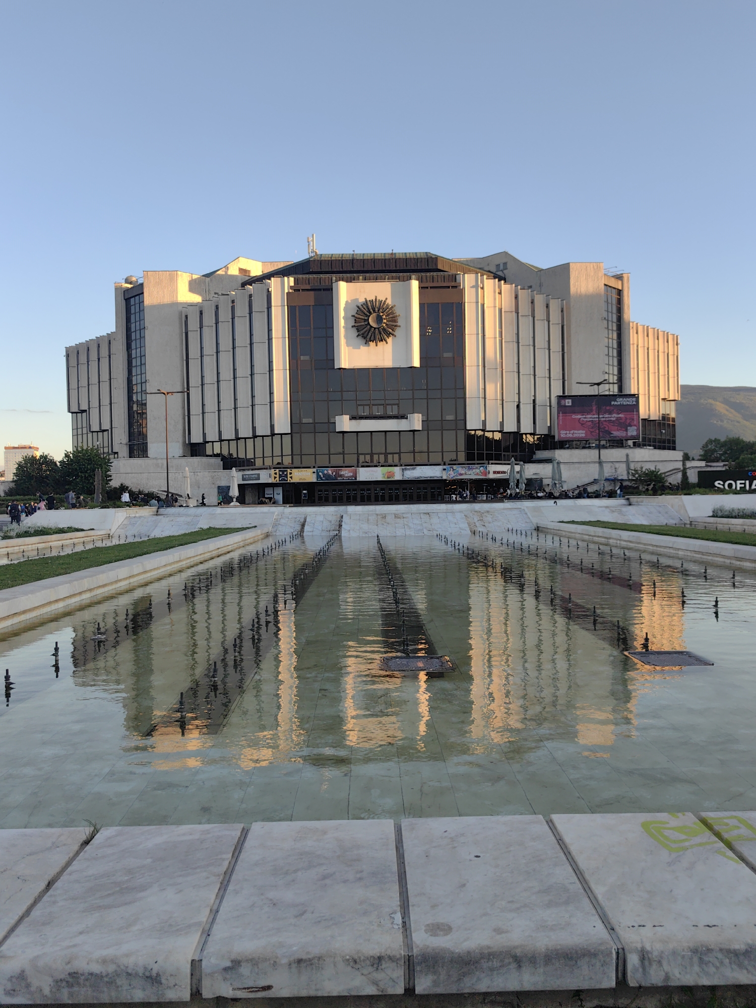 The image depicts the National Palace of Culture in Sofia, Bulgaria, as seen from across a large, rectangular reflecting pool with fountains. The pool occupies a significant portion of the foreground and reflects the building and the late afternoon sunlight. The building is a large, modern structure with prominent vertical window panels and a large, stylized clock or sun decoration on its front facade. To the right of the building is a billboard with promotional material. On the left side of the building, people are gathered on the raised plaza leading to the building's entrance. In the background, a distant mountain range is visible under a clear, blue sky. The overall impression is one of a monumental structure in a public space on a sunny day.