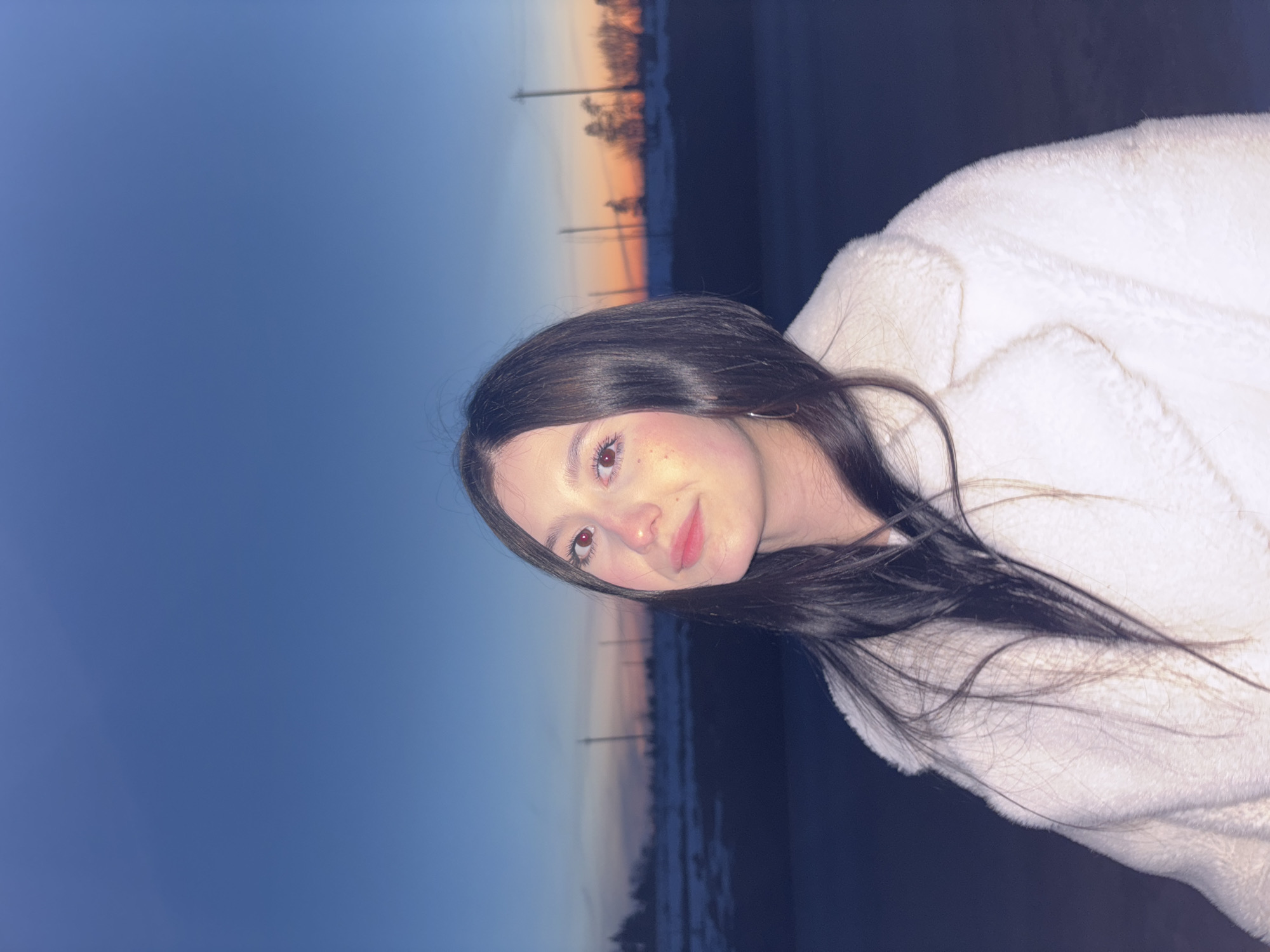 A woman with dark hair and light skin poses for a photograph. She is wearing a white, possibly fur-lined, jacket. The background appears to be a landscape with a gradient sky of blue and orange, possibly indicating sunset or twilight. There are tall, thin structures visible in the background, possibly utility poles. A darker area beneath the sky could be land, water, or a combination of both. It is impossible to locate the image in the city of Фоки, Russia with only the elements visible.