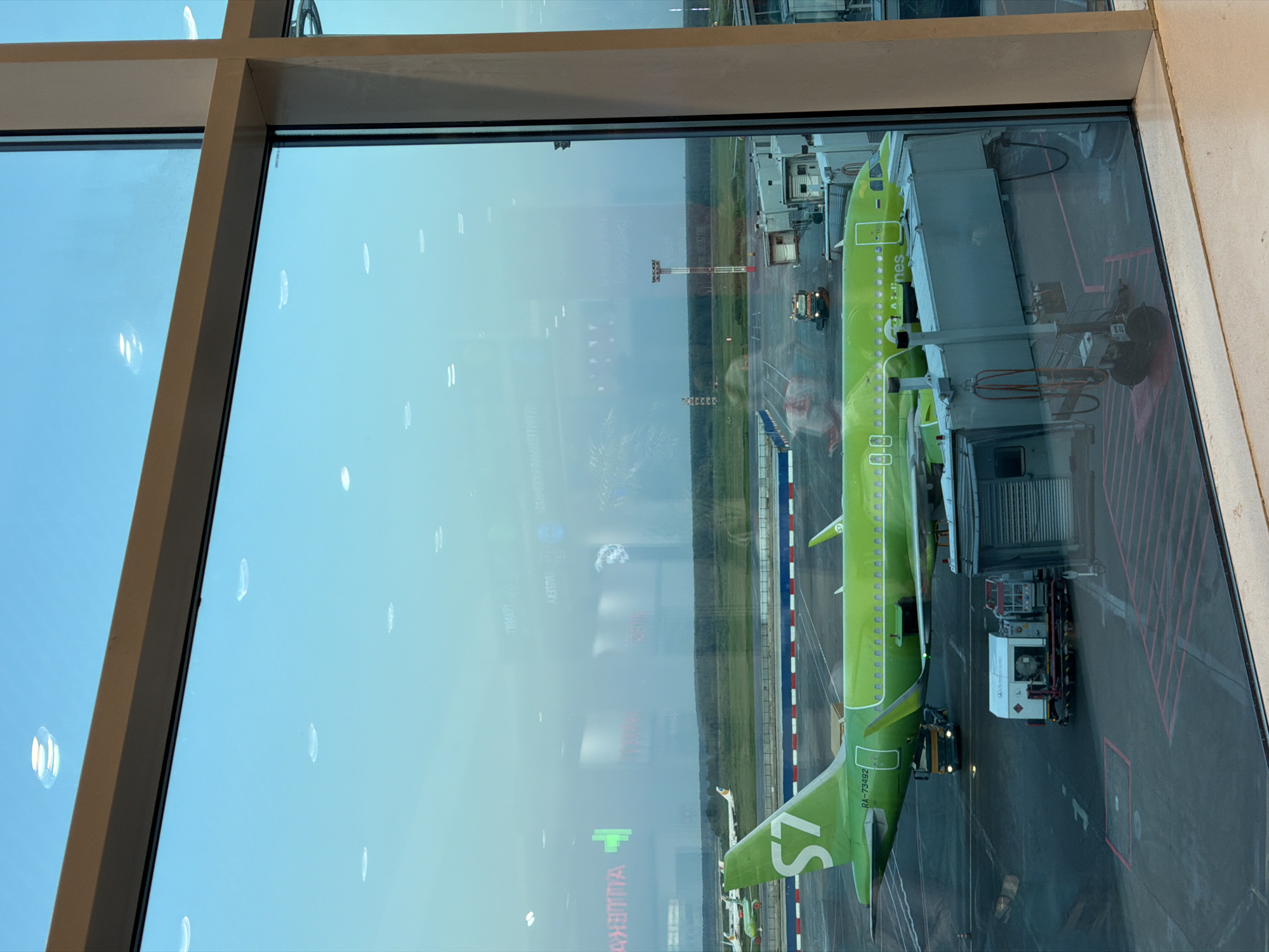 A view through a large window at Domodedovo Airport in Russia shows an S7 Airlines Airbus A320 parked at a gate. The bright green and white livery of the aircraft is prominent. A fuel truck is servicing the plane, with a ground support vehicle nearby. A jet bridge is connected to the aircraft. Other airport infrastructure is visible in the background, including a control tower and more distant buildings. The reflection in the window shows the interior of the airport terminal.