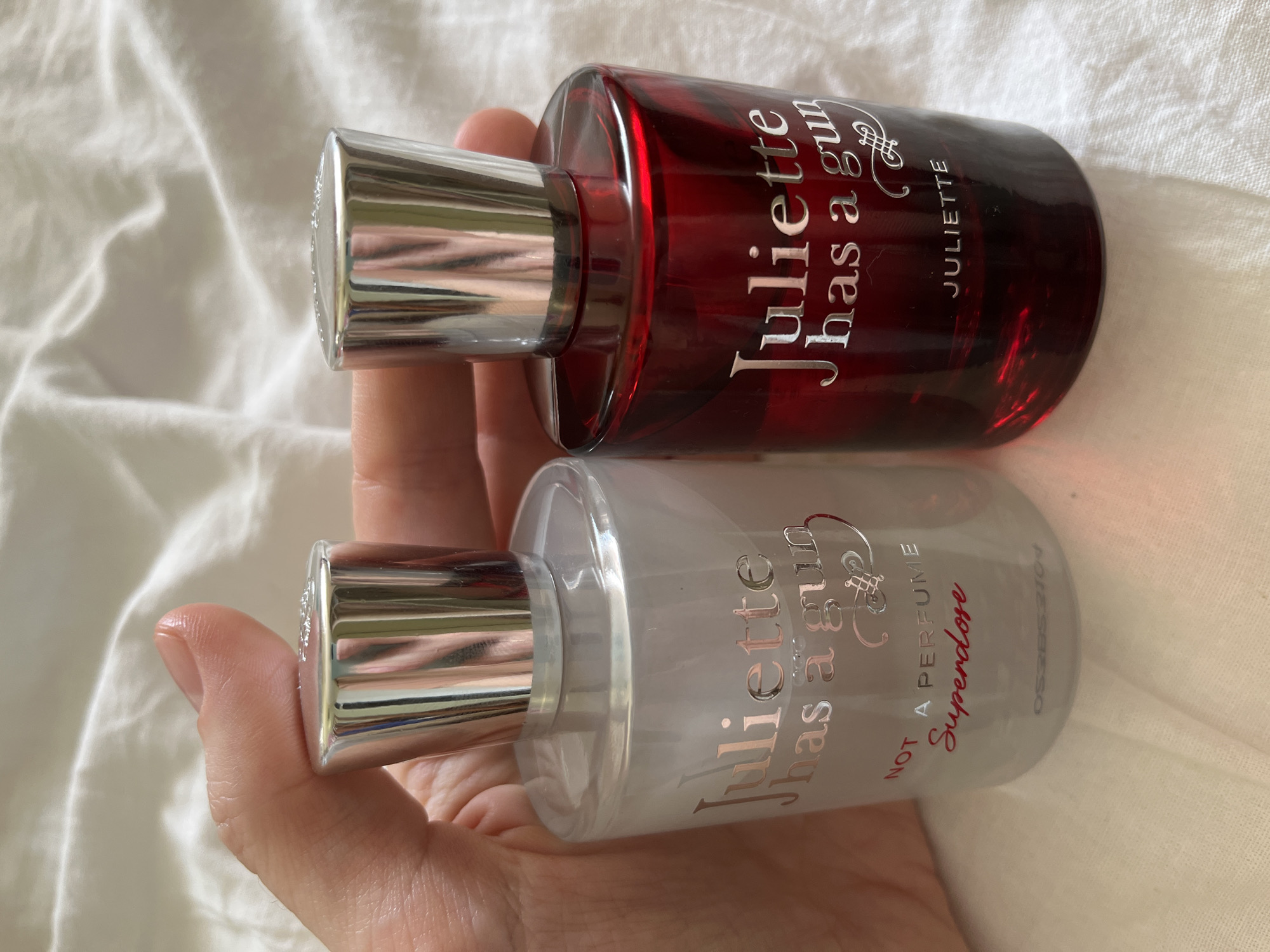 Two bottles of Juliette Has A Gun perfume are held in a person's hand against a white, textured background. The bottle on the left is clear and contains a translucent liquid, with the label "Juliette has a gun" printed in silver, and "NOT A PERFUME Superdose" in red and silver. The bottle on the right is red glass, with the same "Juliette has a gun" label in silver, and "JULIETTE" printed below. The background appears to be a white, wrinkled fabric, possibly bedding. The image does not contain information about the city of Komsomolskoye, Ukraine.