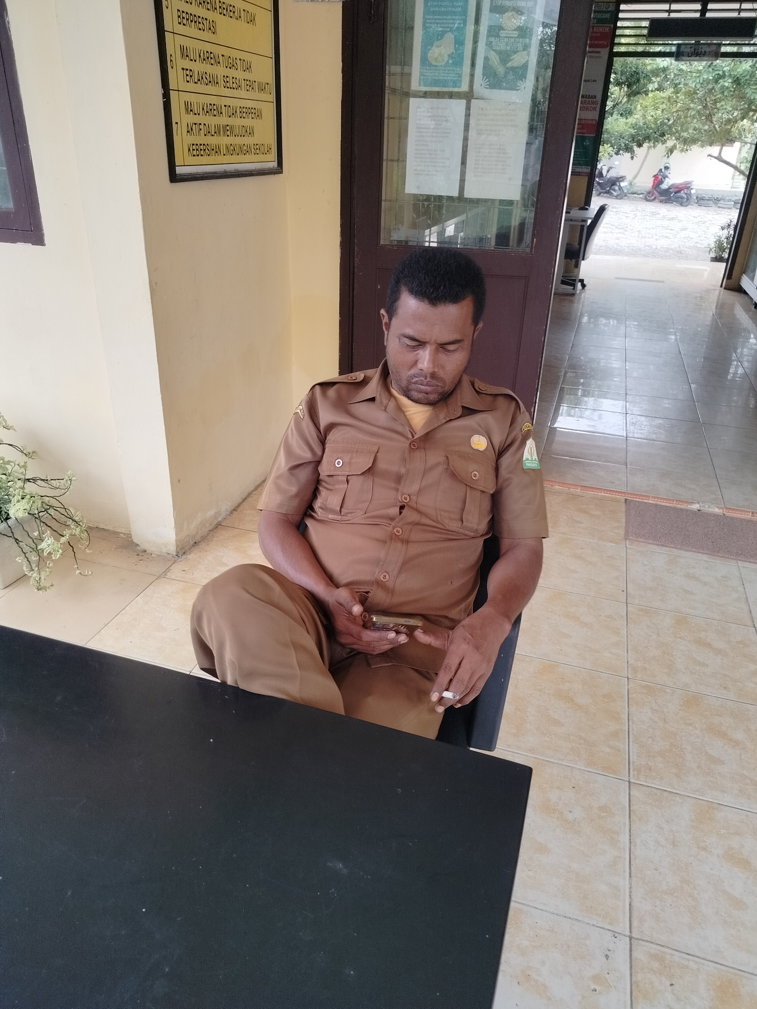 In Lawebekung, Indonesia, a man wearing a brown uniform sits at a black table, looking down at his phone. He holds a lit cigarette in his right hand. The man is seated in a black chair, and behind him, a wall with a framed notice featuring Indonesian text is visible. To his right, a doorway leads to an outdoor area with trees and two motorcycles parked outside. The floor is tiled with light-colored squares. The overall environment appears to be an outdoor or semi-outdoor space, possibly a reception area or waiting space.