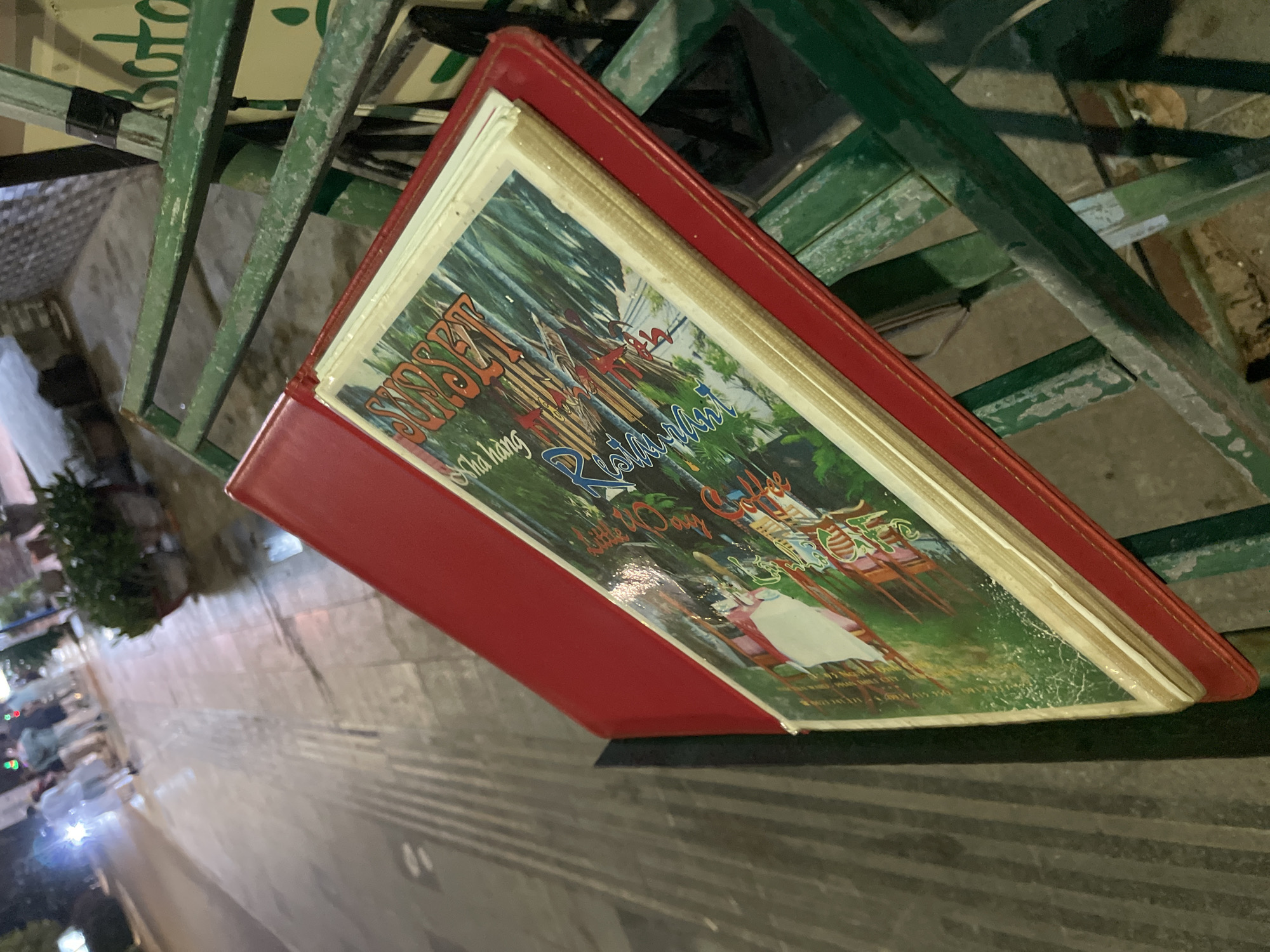 A menu is placed on a green metal rack outside a restaurant in Phu Thuy Ward, Vietnam. The menu has a red cover and a colorful illustration of a tropical scene with palm trees and people. The background shows a street with a few pedestrians and potted plants. The scene is lit by artificial lights, suggesting it is evening or nighttime.
