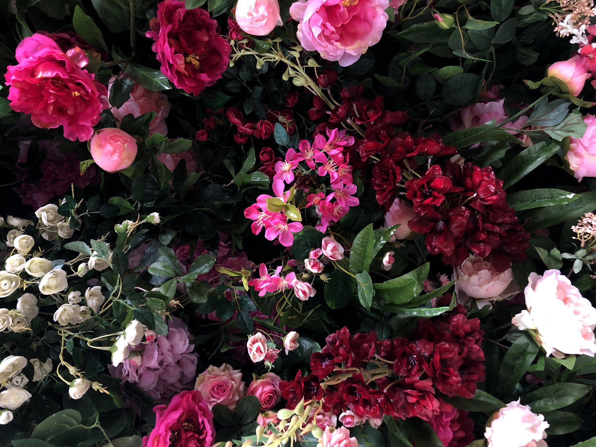 The image depicts a dense arrangement of various flowers and greenery. The flowers are a mix of pink, white, and burgundy hues, with some in full bloom and others in bud. The arrangement includes roses, peonies, and other unidentified floral varieties. Green foliage is interspersed throughout. The photograph appears to be taken in an indoor setting, with artificial lighting contributing to the saturation and contrast. There are no people visible in the frame, focusing primarily on the botanical display. There is no indication of the location of Moscow, Russia.