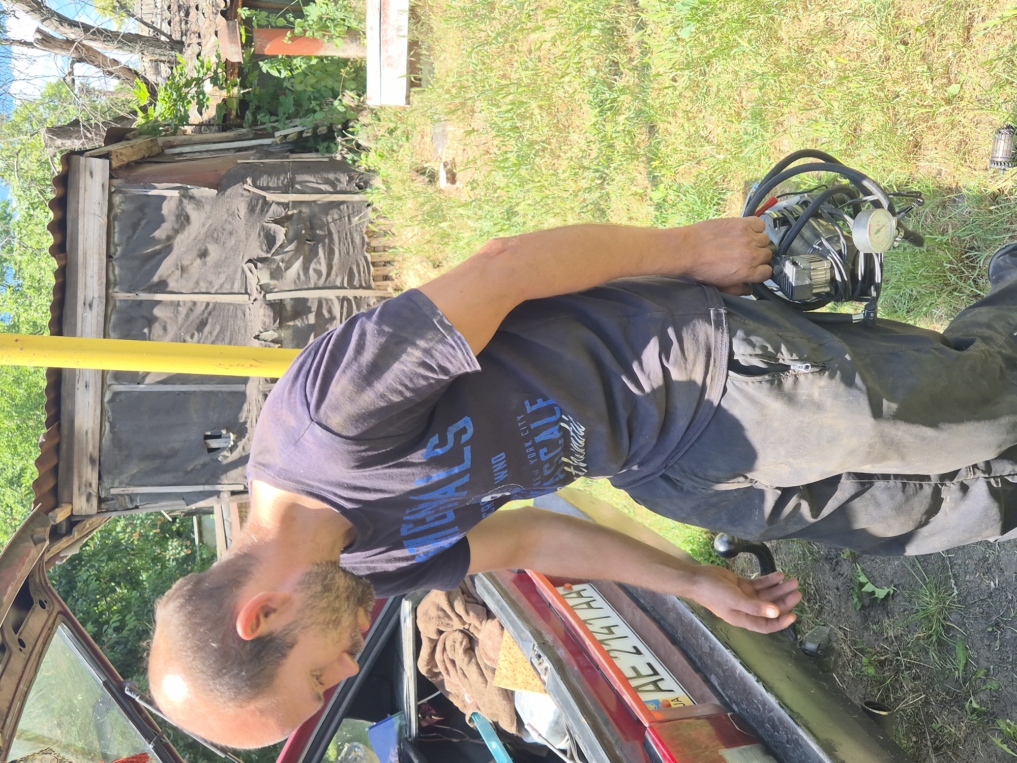 A bald, bearded man wearing a dark t-shirt and dark pants holds a portable air compressor in his right hand. He is standing outdoors in a grassy area next to the open trunk of a red car, which has a license plate with the letters AE 2141 AA. In the background, there is a dilapidated wooden structure with a corrugated metal roof, surrounded by trees and vegetation. The scene suggests a rural or suburban setting.