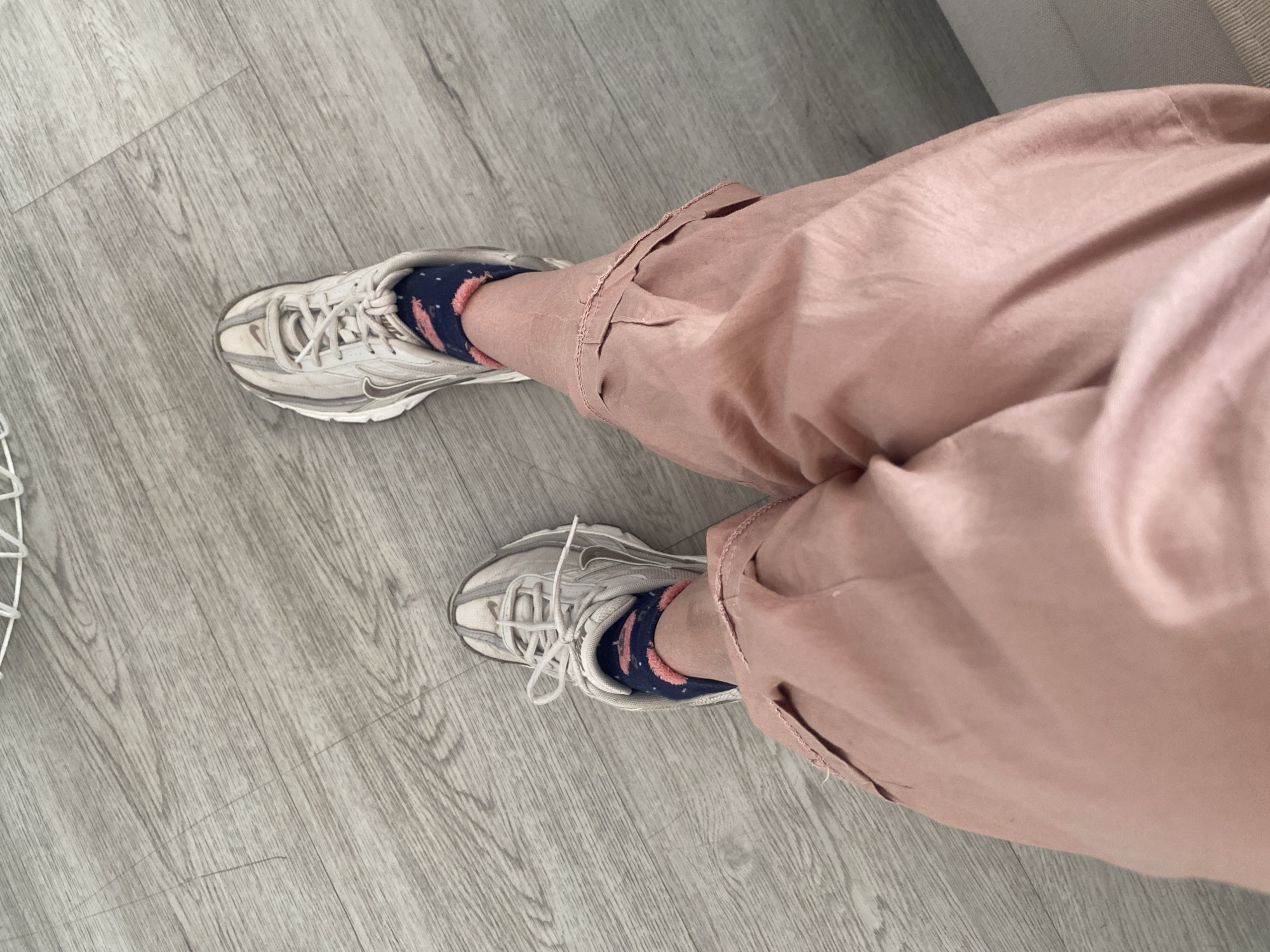 The image shows a person's legs and feet. They are wearing pink cargo pants and white sneakers with blue socks featuring a pink pattern. The person is standing on a gray laminate floor. The location is not specified in the image and is labeled as Unknown, Unknown.