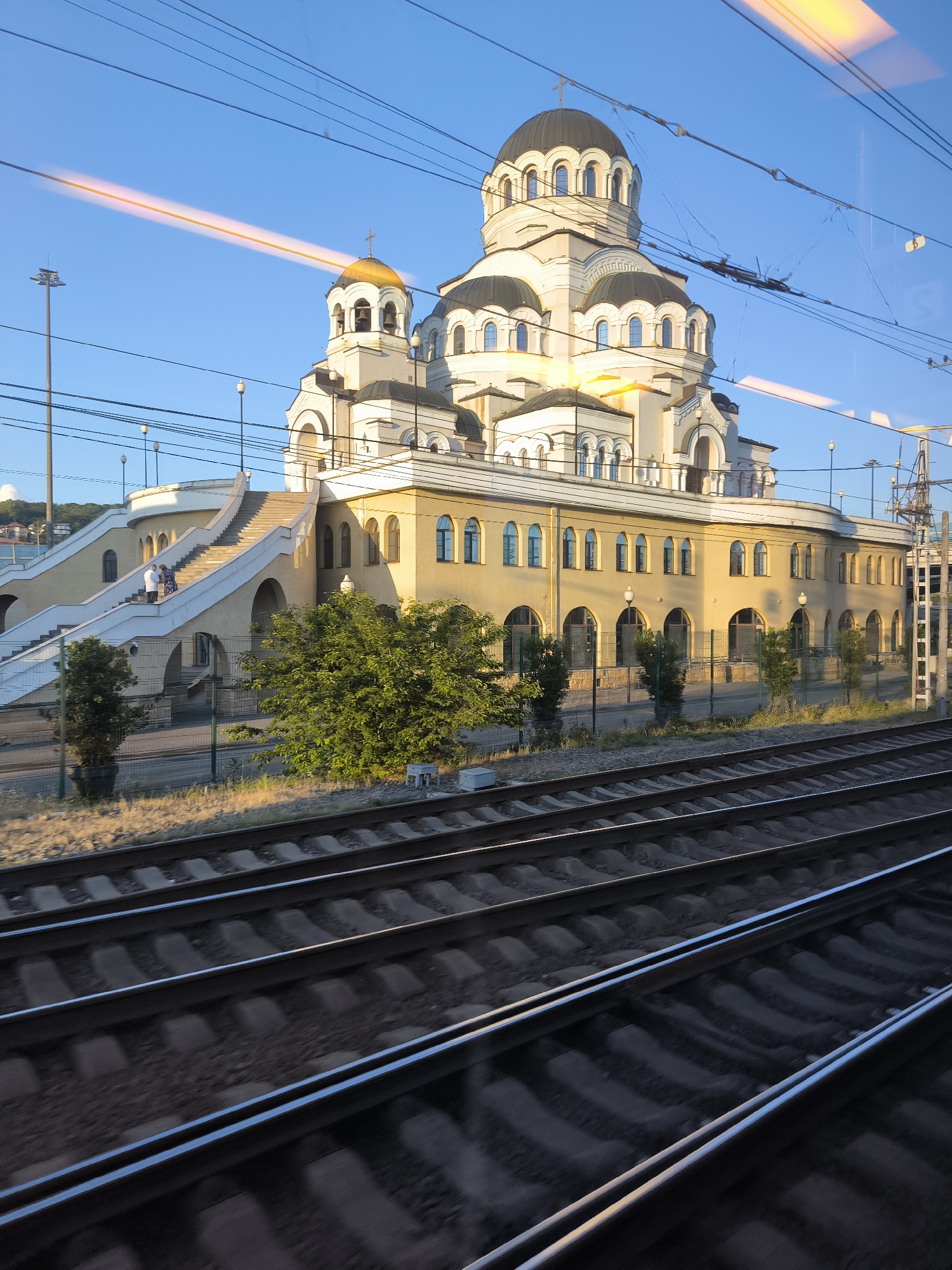 This media file depicts a large, cream-colored building with multiple domes, topped with crosses, suggesting it is a church or cathedral. The building has a grand entrance with a wide staircase leading up to it. Several arched windows line the lower level of the structure. The building is situated in a sunlit environment, indicating it is daytime. In the foreground, there are multiple sets of train tracks, with one set appearing blurred due to motion, implying the photograph was taken from a moving train. A few trees are visible in front of the building, and some people can be seen on the staircase. The sky is a clear blue, with some power lines crisscrossing the upper portion of the image. There are some orange light streaks visible in the sky, possibly reflections or atmospheric effects. The overall scene appears to be serene, with the sunlight casting a warm glow on the building and the surroundings.