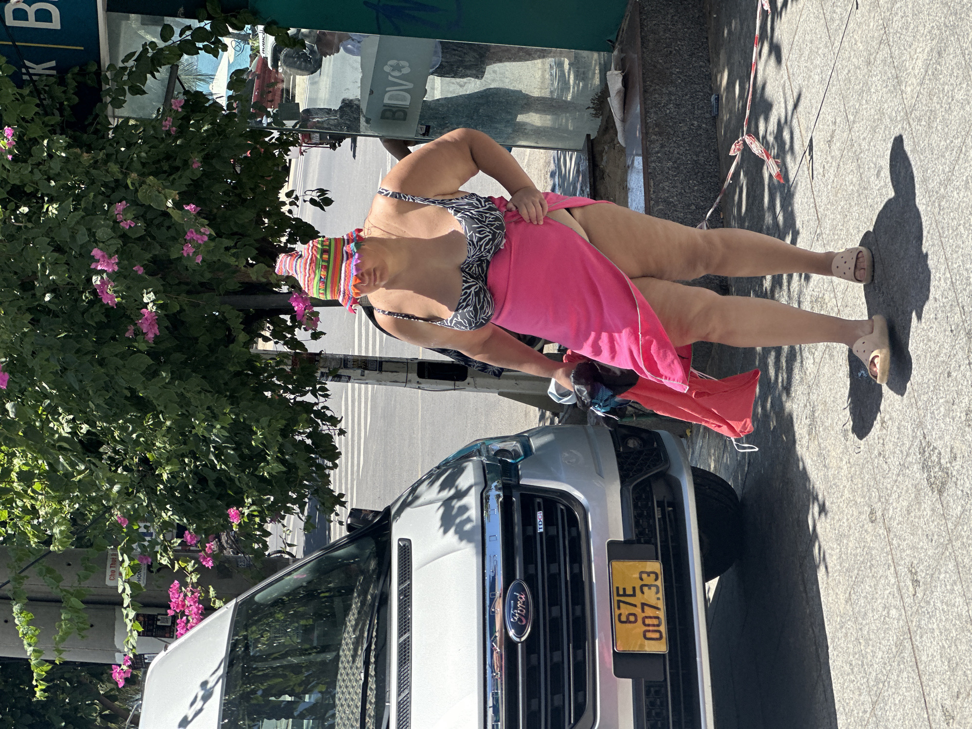 In Bac Nha Trang Ward, Vietnam, a person wearing a colorful, striped hat and a black and white zebra-print bikini top is standing next to a silver Ford van. The person has a pink sarong wrapped around their waist, which is partially lifted, revealing underwear. They are holding a red garment in their left hand. The van has a yellow license plate with the characters "67E 007.33". Behind the person, a glass-fronted building displays the "BIDV" logo. The scene is outdoors, with trees providing shade, and the ground is paved with stone tiles. A construction barrier tape is visible on the ground.