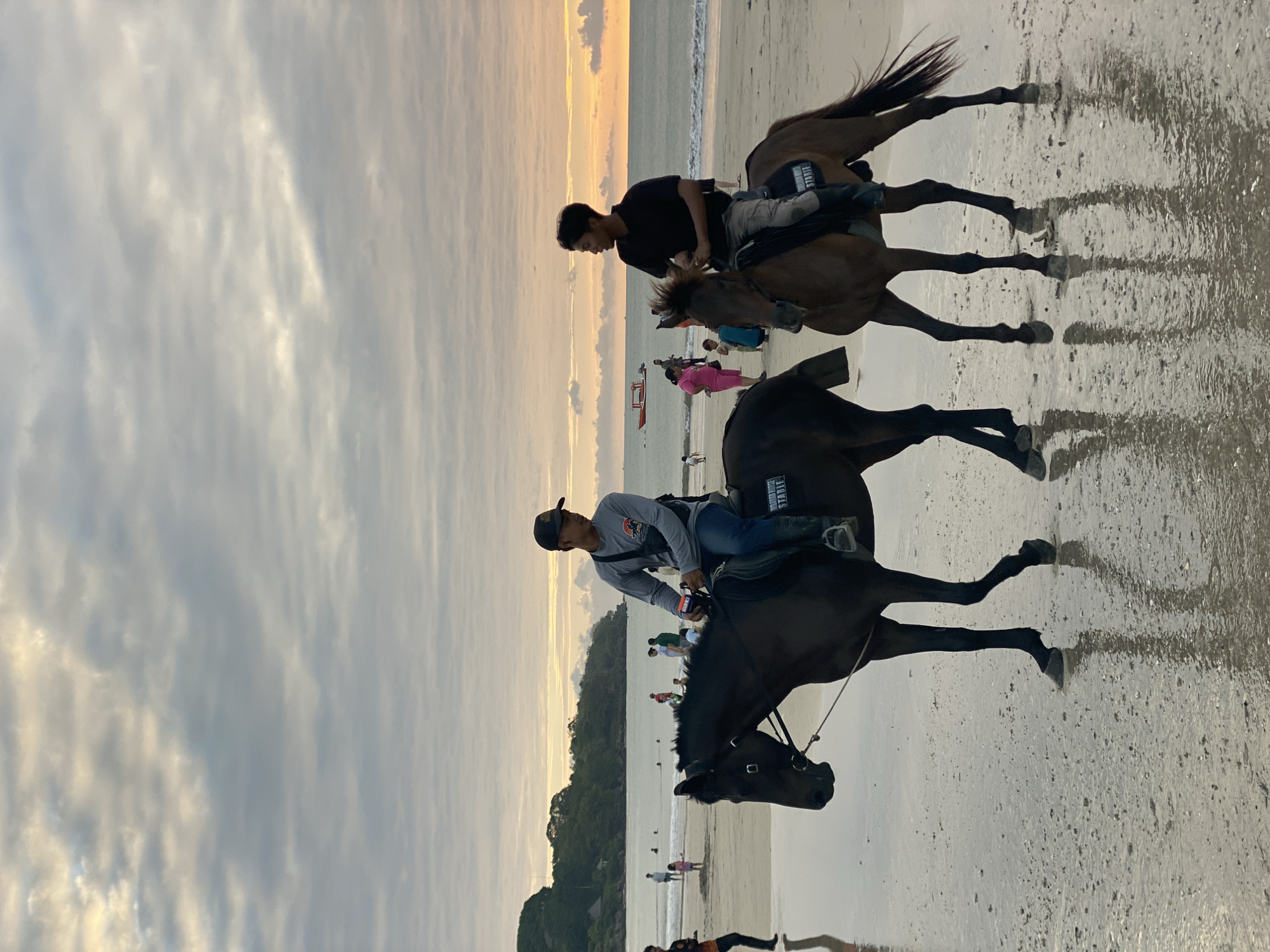 Two men on horseback are walking along the wet sand of a beach in Kuta Selatan, Indonesia. The man on the left is wearing a grey long-sleeved shirt, dark pants, and a black baseball cap, and is riding a dark brown horse. The man on the right is wearing a black t-shirt and dark pants, and is riding a chestnut horse. Both horses have saddles and bridles. The horses' hooves are making imprints in the wet sand, and their reflections are visible on the wet surface. In the background, the ocean stretches out to the horizon, with a few boats visible. The sky is cloudy, with hints of orange and yellow from the setting sun. Other beachgoers are scattered in the background, some walking, some in the water. A lush green hill is visible on the left side of the image. The overall scene depicts a peaceful moment at the end of the day on a popular Indonesian beach.