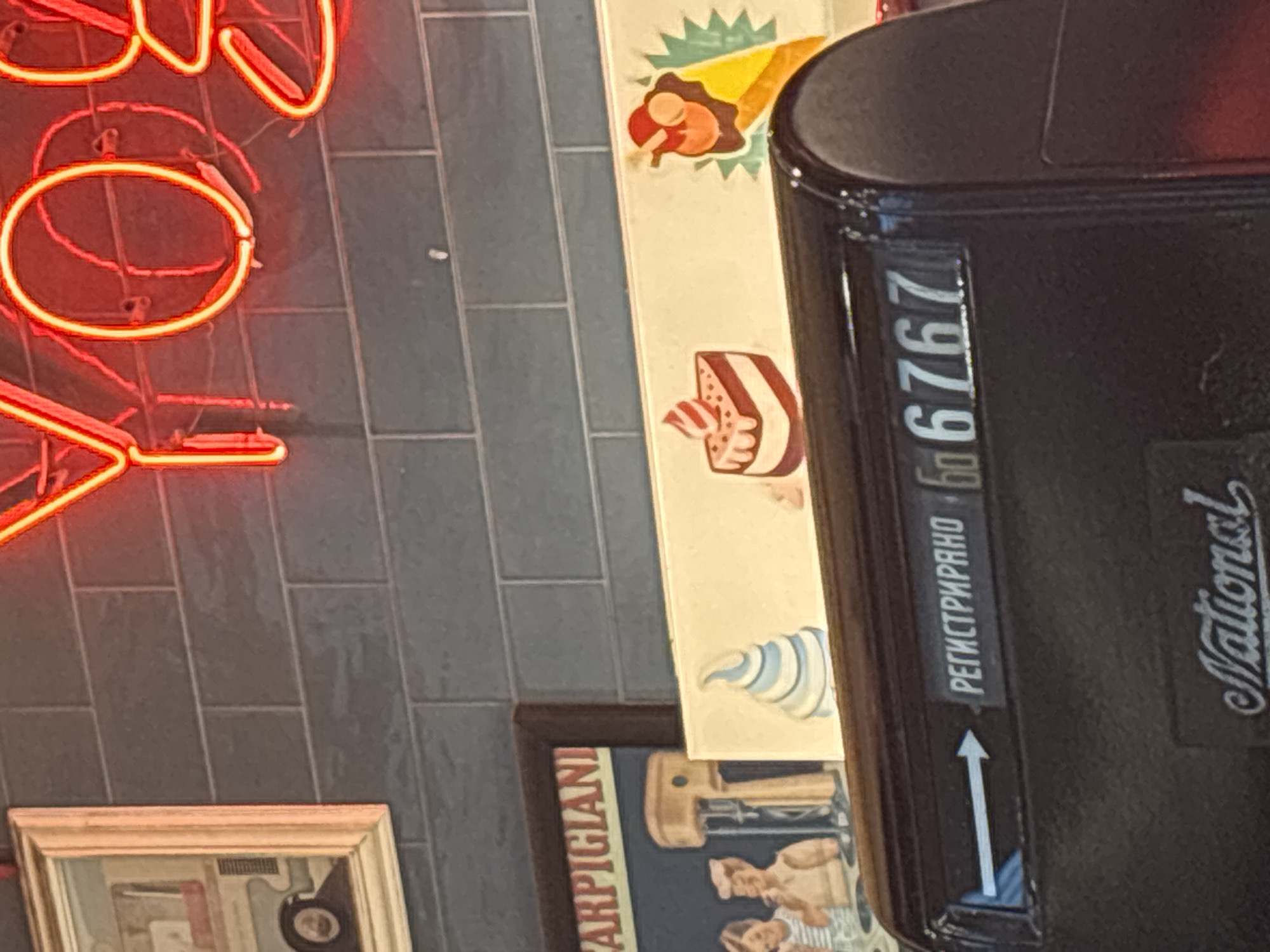 The image captures an indoor scene, likely in a commercial establishment such as a restaurant or cafe, featuring various decorative elements. On the left, a bright red neon sign, partially visible, displays the glowing outlines of letters including a clear "O." The wall behind it is composed of dark grey, rectangular tiles.
To the right, a vertical cream-colored panel is decorated with colorful illustrations of food items. These include a dessert in a cone with a green starburst, a slice of layered cake, and a blue and white striped cone-shaped item.
A prominent black, glossy object, possibly part of an antique cash register or counter, occupies the right side of the frame. It features an upward-pointing arrow and text in Cyrillic script: "РЕГИСТРИРАНО" (Registrirano, meaning "Registered"). Below this, "ба 6767" is visible, likely a numerical counter or serial number. The brand "National" is also displayed on the side of this object.
At the bottom left, a corner of a framed picture or artwork is visible against the tiled wall. Text that appears to be "PARPIGIANI" (possibly "Parmigiani") can be faintly read on it, along with illustrations of figures and what looks like a kitchen tool. The scene is static, depicting interior decor without any people or specific activities.