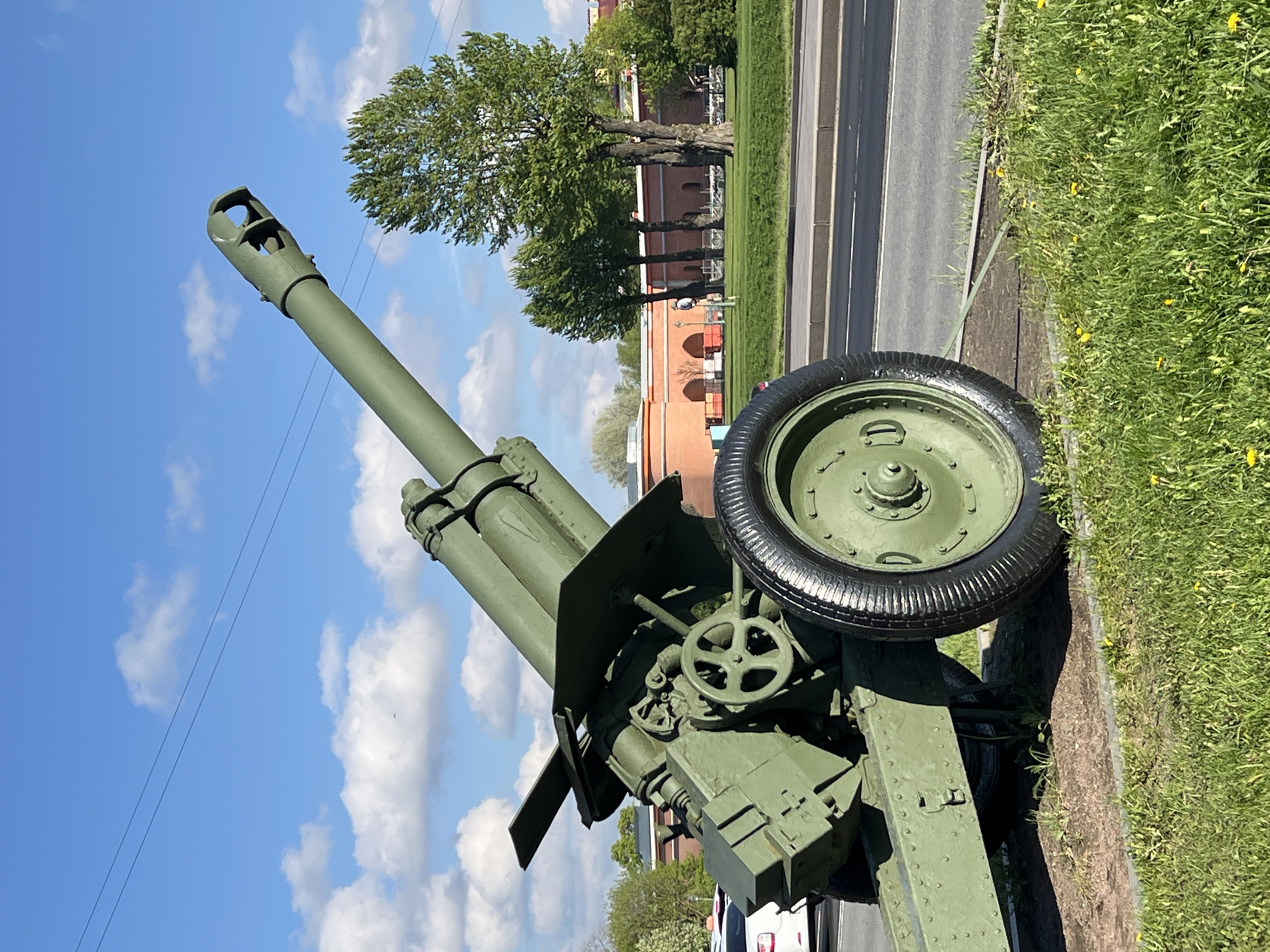 The image shows a green artillery gun in Saint Petersburg, Russia. The gun is positioned on a grassy area adjacent to a road and in front of a red brick building with arched windows. The sky is blue with scattered white clouds. There are trees in the background. A tire is mounted on the side of the gun. A white vehicle is partially visible at the bottom of the frame.