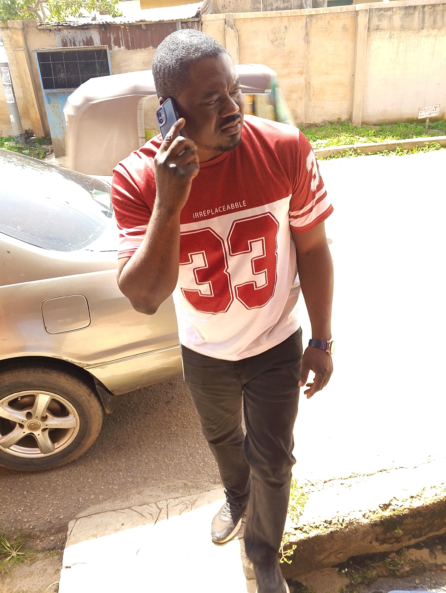 A man, appearing to be in his 40s with dark skin and a short, buzzed haircut, stands on a paved area outdoors, talking on his dark-colored smartphone held to his right ear. He is wearing a red and white t-shirt with the number "33" prominently displayed in red on a white background, and the word "IRREPLACEABLE" in small white text above it. He is also wearing dark-colored trousers and grey athletic shoes. On his left wrist, he has a wristwatch with a dark band and a circular face.
To his left, a silver sedan is parked, with its rear wheel and part of the door visible. The car appears to be in an urban or semi-urban environment, indicated by the paved road and a concrete curb with some vegetation growing alongside it. In the background, there is a building with a weathered concrete wall, and further back, a structure with corrugated metal roofing and a blue door with a barred window. A small, three-wheeled vehicle, possibly a commercial tricycle, is partially visible behind the man and the sedan.
The lighting suggests it is daytime, likely mid-morning or early afternoon, with bright sunlight casting shadows. The overall scene conveys a casual, everyday moment. No explicit information about the weather is available, but the bright light suggests it is likely sunny. The presence of the car and the buildings indicates an urban or suburban setting, consistent with the provided location context of Jos, Nigeria.