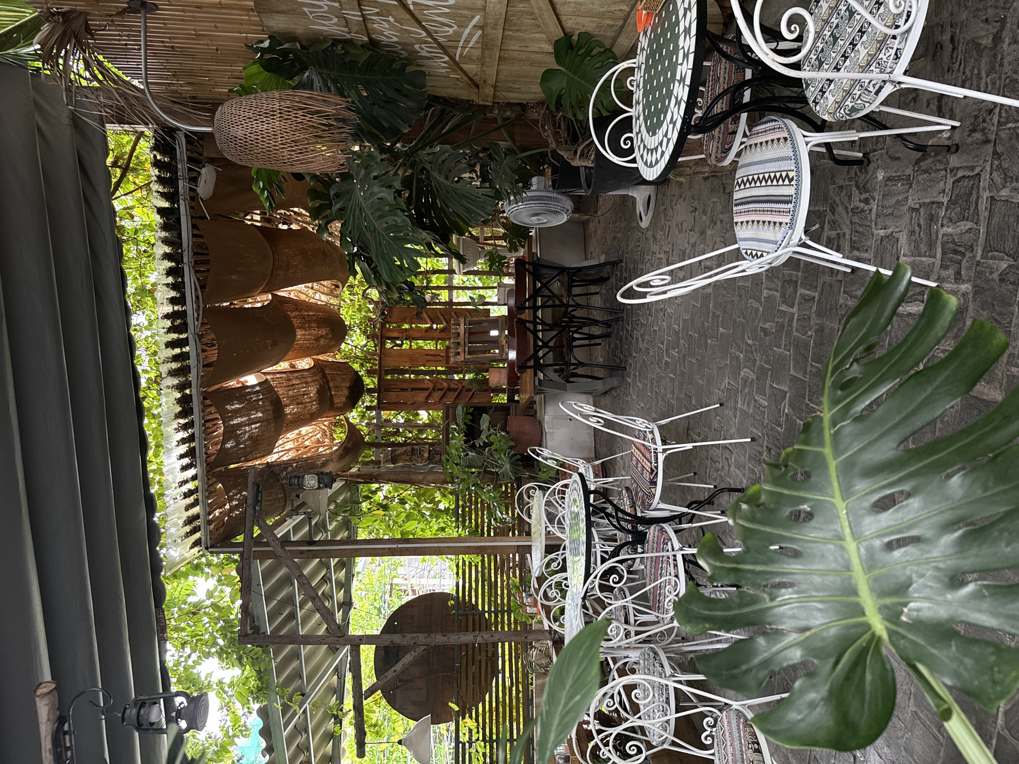 The image shows an outdoor seating area at a restaurant or cafe in Nha Trang, Vietnam. Several round tables with mosaic tops and white wrought-iron chairs with patterned cushions are arranged on a stone-tiled floor. Lush green foliage, including large Monstera leaves, hangs from above and surrounds the seating area, creating a natural, tropical ambiance. In the background, wooden structures, bamboo accents, and woven light fixtures contribute to the rustic decor. A white oscillating fan stands near a large potted plant. The overall scene suggests a relaxed and casual dining or gathering space.