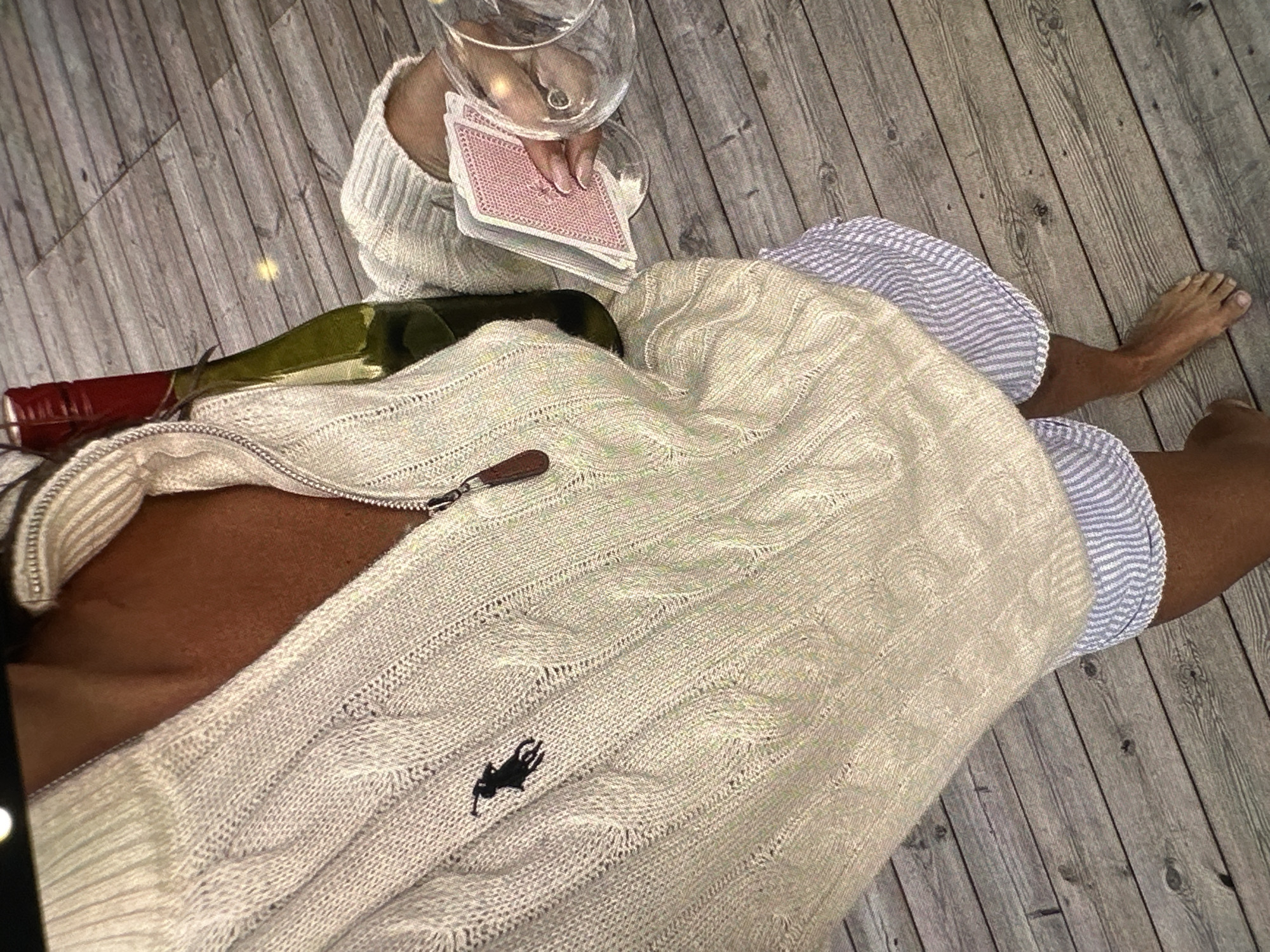 A person is sitting on a grey wooden floor, wearing a partially zipped, light-colored cable-knit sweater with a black polo emblem, and striped shorts. A wine bottle lies on the sweater, and a hand holding a stack of playing cards is visible near a wine glass. The location is unknown, not necessarily in Yerevan, Armenia.