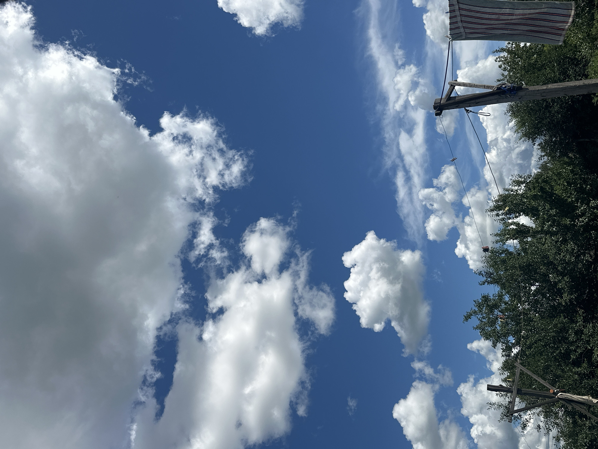 The image shows a clear blue sky with scattered white clouds. In the foreground, there is a line of trees. A wooden post with some items hanging on a wire is visible on the right side of the image. One item appears to be a striped towel. There are no people in the image. The location is identified as the city of Tobyl, Kazakhstan.