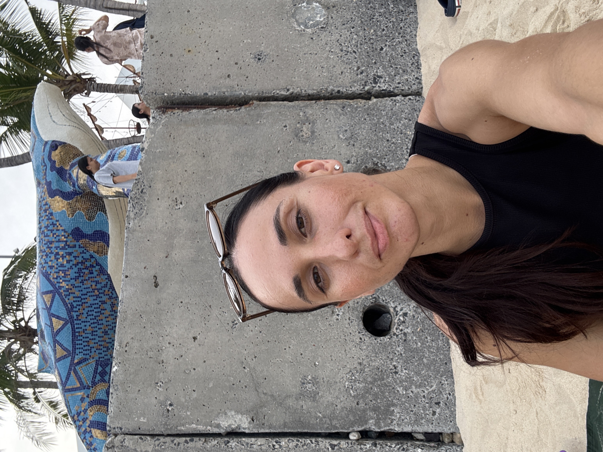 A woman wearing a black top and glasses is posing in front of a concrete structure. In the background, there is a blue and gold mosaic statue of a whale or sea creature near palm trees, suggesting a beach or coastal area. There are other people in the background, though they are not the main focus. The location is Nha Trang Ward, Vietnam based on similar locations.