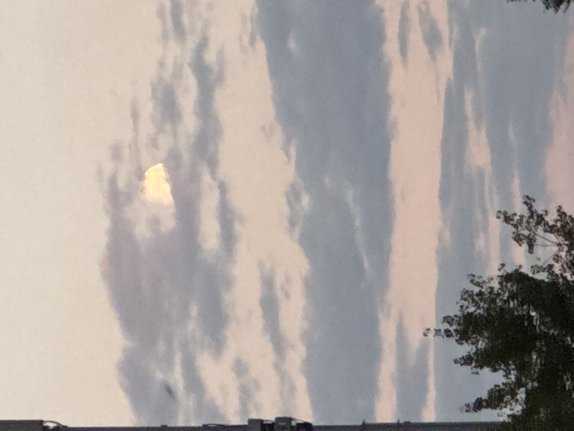 A cell tower is visible on the left side of the frame, partially obscuring the view of a cloudy sky during sunset or sunrise. The sky displays soft hues of pink and orange, with scattered gray clouds. Partially visible through the clouds is a luminous crescent moon, emitting a soft yellow glow. At the bottom of the image, the dark silhouettes of trees are present, suggesting a natural or park-like setting within or on the outskirts of Ufa, Russia.