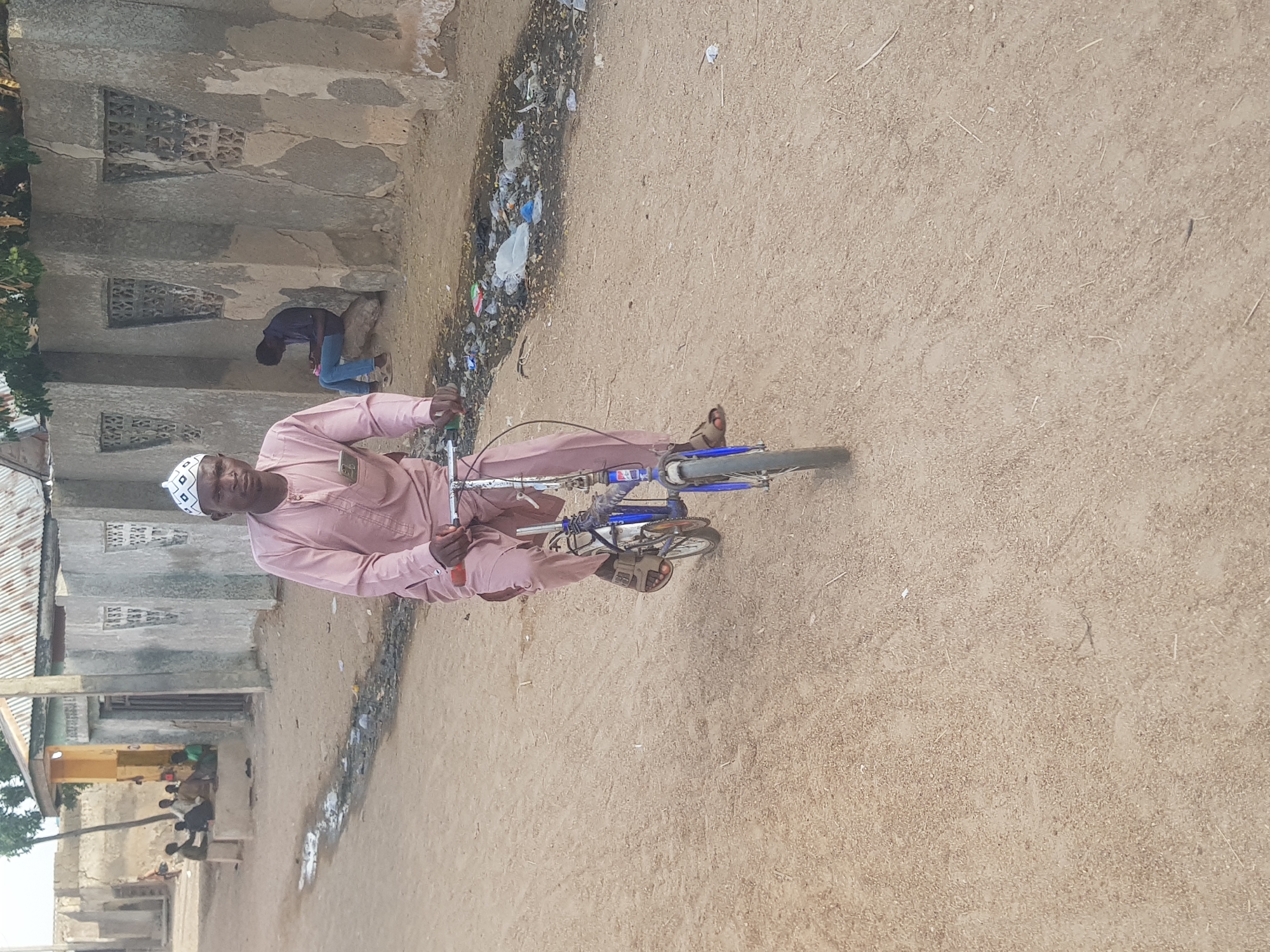 A man is riding a bicycle on a dirt road in Dutsinma B, Nigeria. He is wearing a light pink, long-sleeved shirt and pants with a white and black kufi. A blue bicycle is underneath him. The dirt road is light tan in color, and littered with light debris. Behind him, to his left, is a light gray concrete building with small, decorative window openings. A small group of people is visible at the side of the building. To his right, there is a ditch with trash, primarily plastic, visible in the foreground. The weather appears to be sunny and bright.