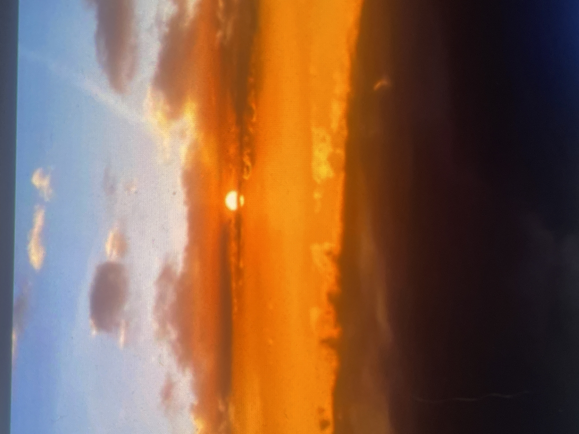 The image depicts a sunset or sunrise over Zelenyi Hai, Ukraine. The upper portion of the frame features a light blue sky with scattered white clouds, one of which appears distinctly rectangular in the upper-left quadrant. A faint diagonal light streak is visible in the upper-right sky. Slightly left of the image's vertical center, a distinct, circular, bright orange sun is partially obscured by a dense layer of vivid orange-red clouds that extend horizontally across the mid-section. Below this bright orange band, the scene transitions into a dark, deep brown to black mass, likely representing shadowed terrain or low-lying clouds, where specific features are largely indistinguishable. A faint, slender, light-colored vertical anomaly is present near the bottom-left edge, and a small, curved luminous artifact is visible in the bottom-right within the dark foreground. No human figures or discernible man-made structures are present. The image exhibits a visible pixel grid pattern and other minor artifacts, suggesting it is a photograph of a digital display rather than a direct capture.