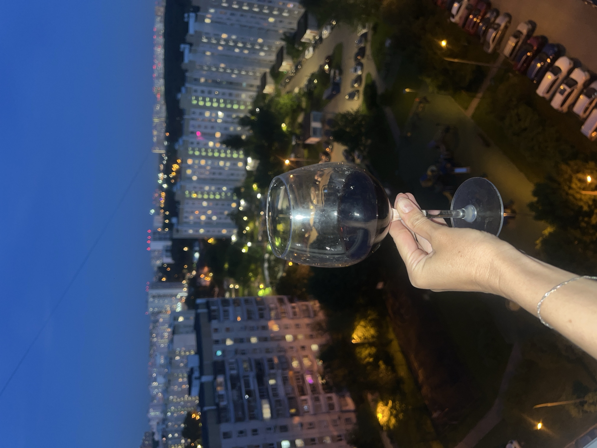 A glass of red wine is held in front of a cityscape at night. The hand holding the wine glass appears to be Caucasian with a silver bracelet on the wrist. The cityscape is Moscow, Russia. High-rise buildings with multiple illuminated windows, indicative of residential apartments, dominate the background. Below the buildings are streetlights, a parking lot with cars, and a playground. The sky is dusky blue, suggesting dusk or early evening.