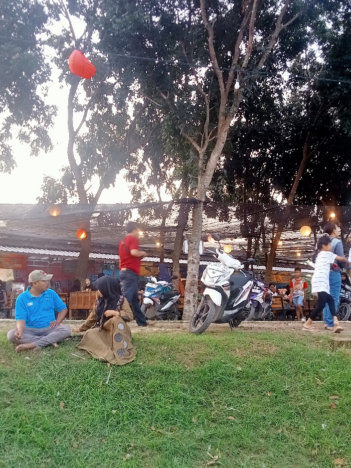 The image shows a scene in Jakenan, Indonesia, located at coordinates -6.758 latitude and 111.075 longitude. The scene takes place in a grassy area next to a restaurant or cafe. Three adults and two children are present. Two adults are sitting on the ground, one man wearing a blue shirt and the other woman wearing a brown hijab and a long coat. Another man is standing, wearing a red shirt, and there is a woman and child walking to the left. The woman is wearing a white and blue dress and the child is wearing a pink dress. In the background, a group of people is sitting at a table. There are three motorcycles parked behind the group, and two more motorcycles can be seen in the background. The cafe has a wooden roof and is covered by a dark canopy. Strings of light bulbs hang from the canopy, and there are other light bulbs hanging from the trees. A red balloon is floating in the sky. The ground is covered with green grass and a few white rocks. The image was likely taken in the late afternoon or early evening, as the sun is low in the sky and casting long shadows. The overall mood of the image is relaxed and peaceful.