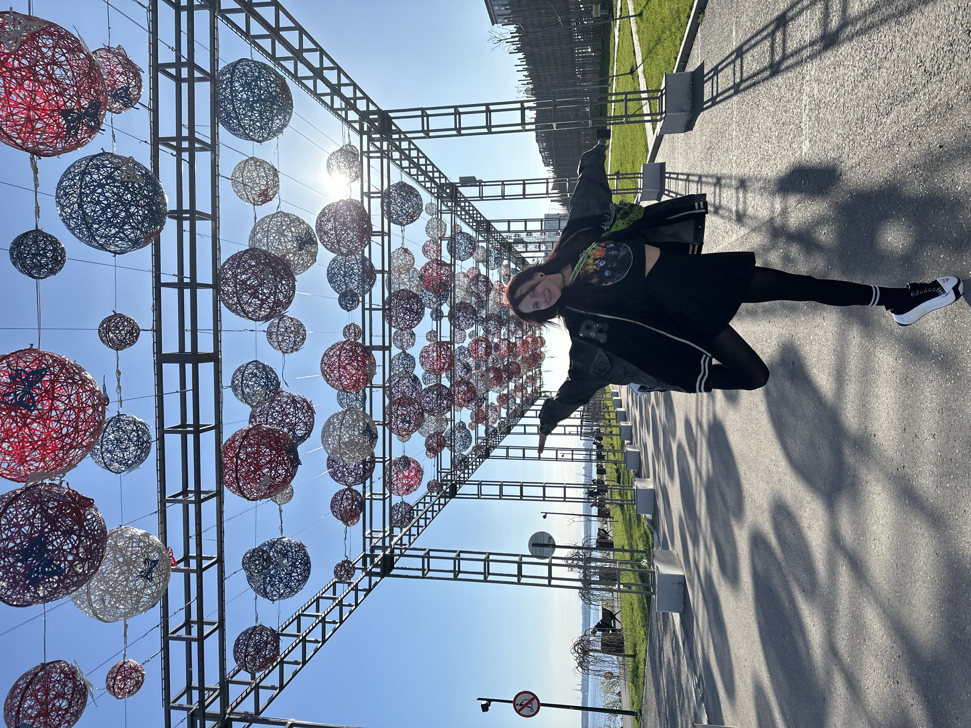 The image captures a vibrant outdoor scene on a sunny day in Saratov, Russia. A young person, likely female, with dark hair, is depicted in an energetic pose, seemingly mid-jump, with arms outstretched and a joyful expression. She is dressed in a black jacket, a dark top with a graphic print, a black skirt, dark tights, and black and white platform sneakers.
The setting is a paved walkway or path, illuminated by bright sunlight. Above the path, an extensive metal truss structure supports numerous decorative, spherical, open-weave balls. These balls are predominantly red, white, and blue, creating a festive overhead display. The sun, visible in the upper right quadrant, casts long, defined shadows of the structure and the hanging balls onto the paved ground, indicating a clear day, likely mid-morning or late afternoon.
To the right of the walkway, there is a stretch of green grass and some trees. Beyond this, a large body of water, possibly the Volga River, extends towards the horizon, with distant land or city structures faintly visible. Benches and concrete bollards line the pathway. A red and white "no parking" sign is visible in the lower right foreground. The scene suggests a lively public park or waterfront promenade.