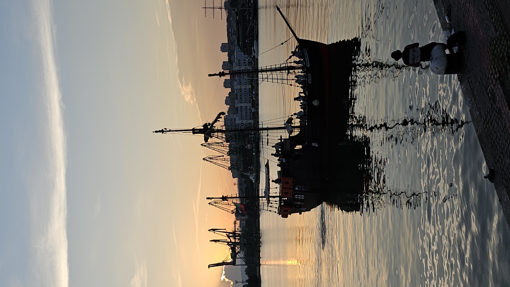 This is a vertical, medium-shot, silhouette photograph of a pirate ship sailing on the sea in Varna, Bulgaria, during sunset. The ship is in the foreground and is silhouetted against the orange and yellow sky. The ship is dark and has two masts. There are people on the deck of the ship. In the background, there are cranes and buildings. The sun is setting behind the cranes. The water is calm and reflects the sky and the ship. A person wearing a hat is sitting on a bollard in the lower right corner of the image.